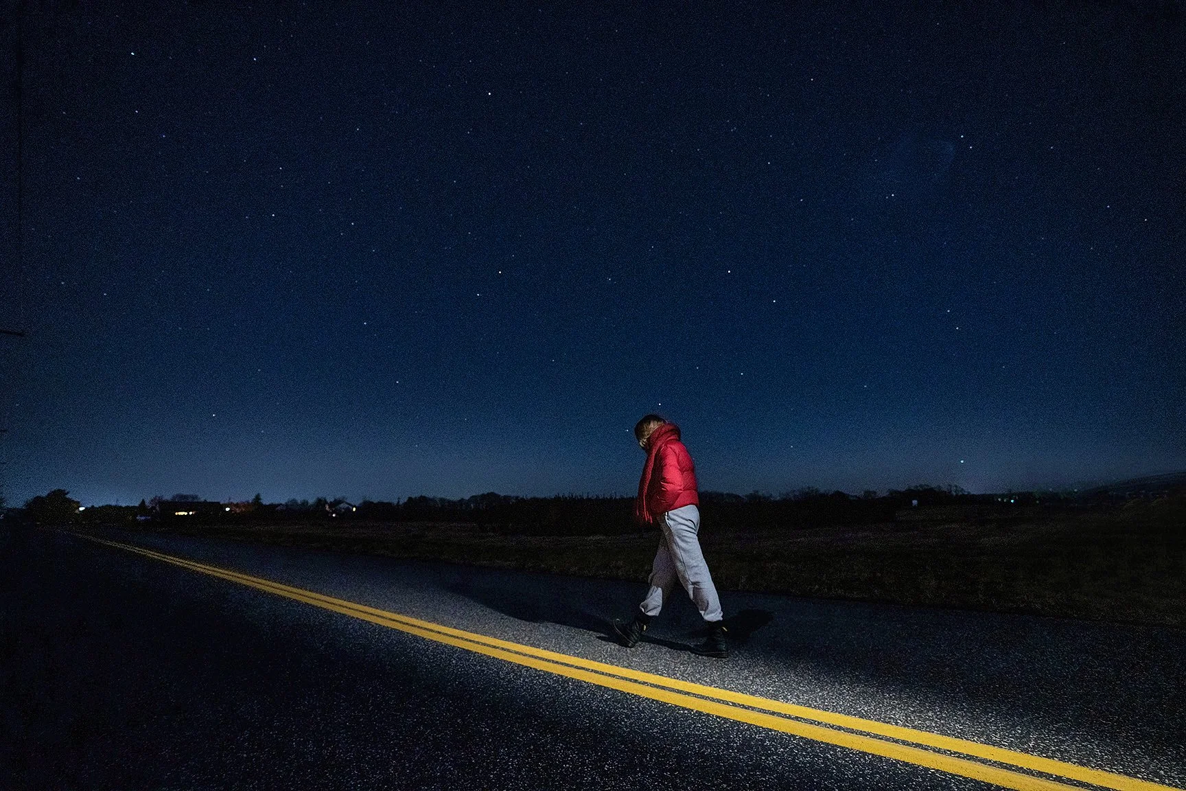 A person walking along a dark road at night, wearing a red jacket and gray pants, under a starry sky.