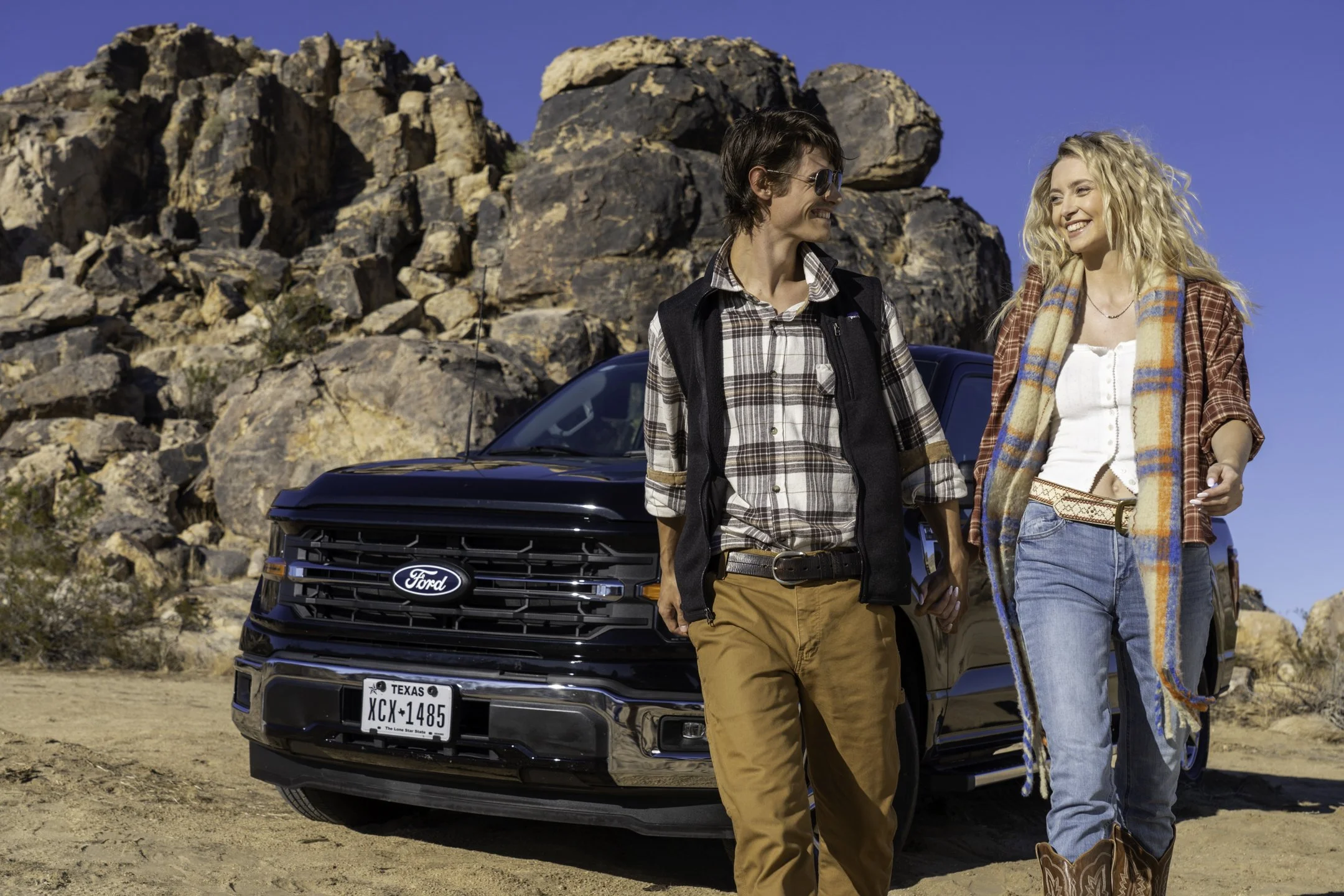 Two people, a man and a woman, are walking in a desert area with large rocks behind a black Ford vehicle. They are smiling and looking at each other. The man has short dark hair, sunglasses, a plaid shirt, and khaki pants. The woman has long curly bl
