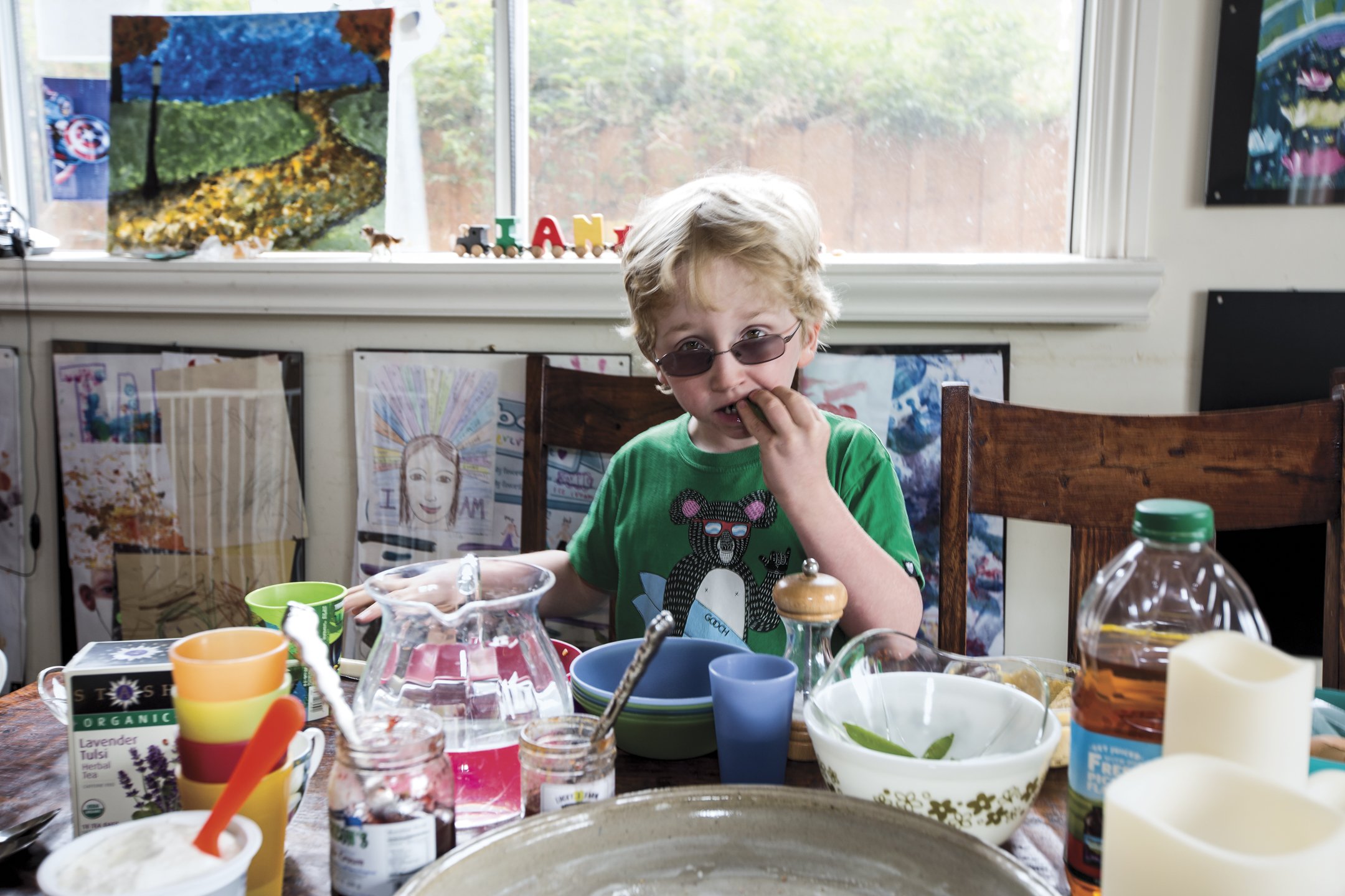 A young boy with curly blond hair wearing sunglasses and a green T-shirt, sitting at a dining table filled with various dishes and drinks, during daytime in a room with artworks and drawings on the wall and colorful decorations on the window.