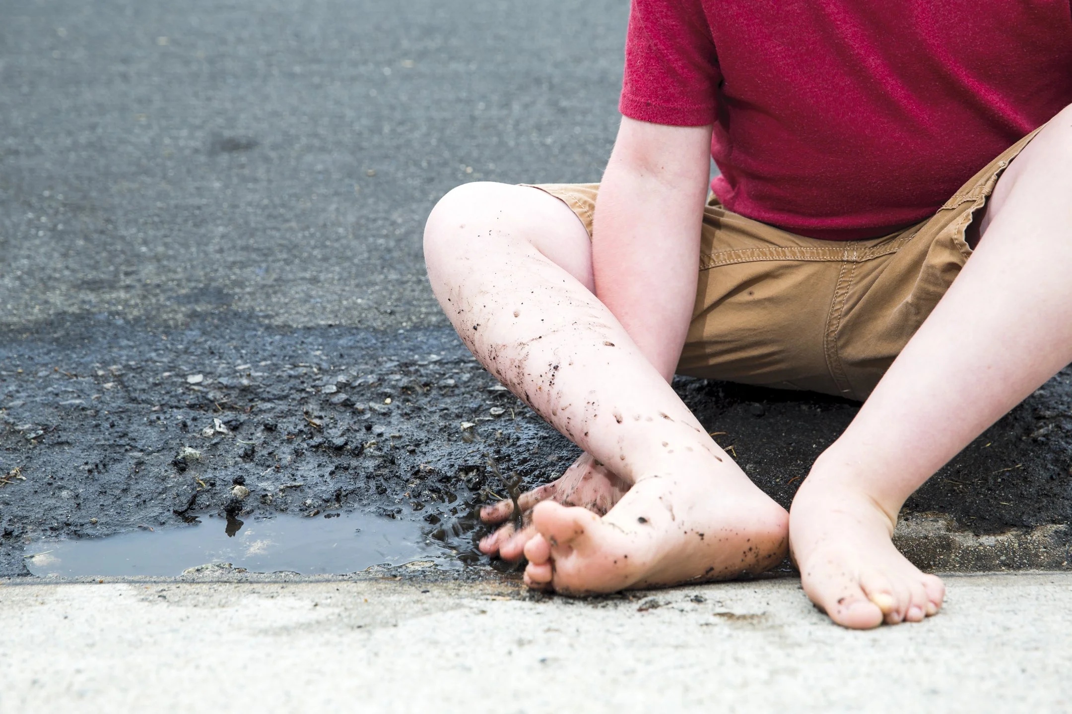Child sitting on the side of a road with muddy feet, near a puddle of water, wearing a red shirt and tan shorts.