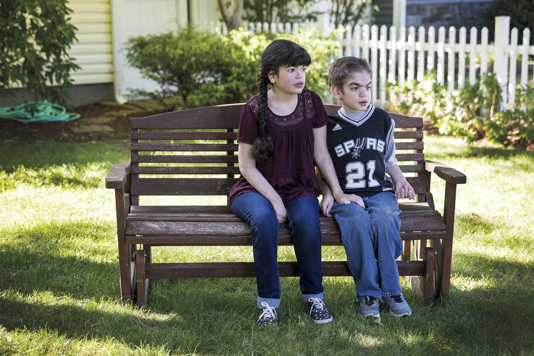 A boy and girl sitting on a wooden bench in a backyard, looking to the side with serious expressions, surrounded by green grass, bushes, and a white picket fence.