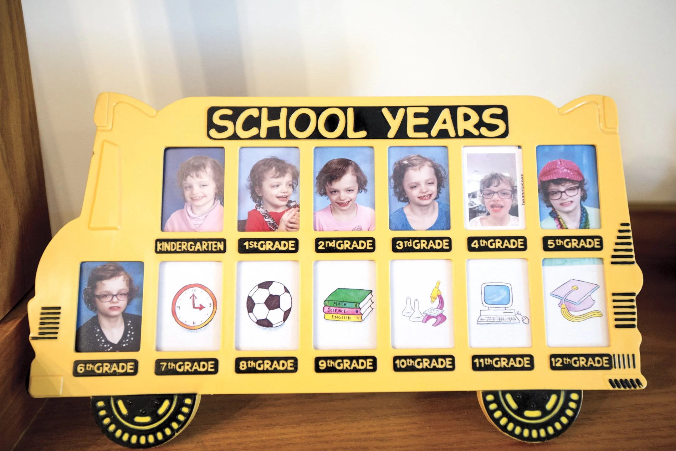 A yellow school bus-shaped display board with photographs of a girl at different ages from kindergarten to 12th grade. Each grade is labeled beneath the photos, with some images including drawings representing school subjects.