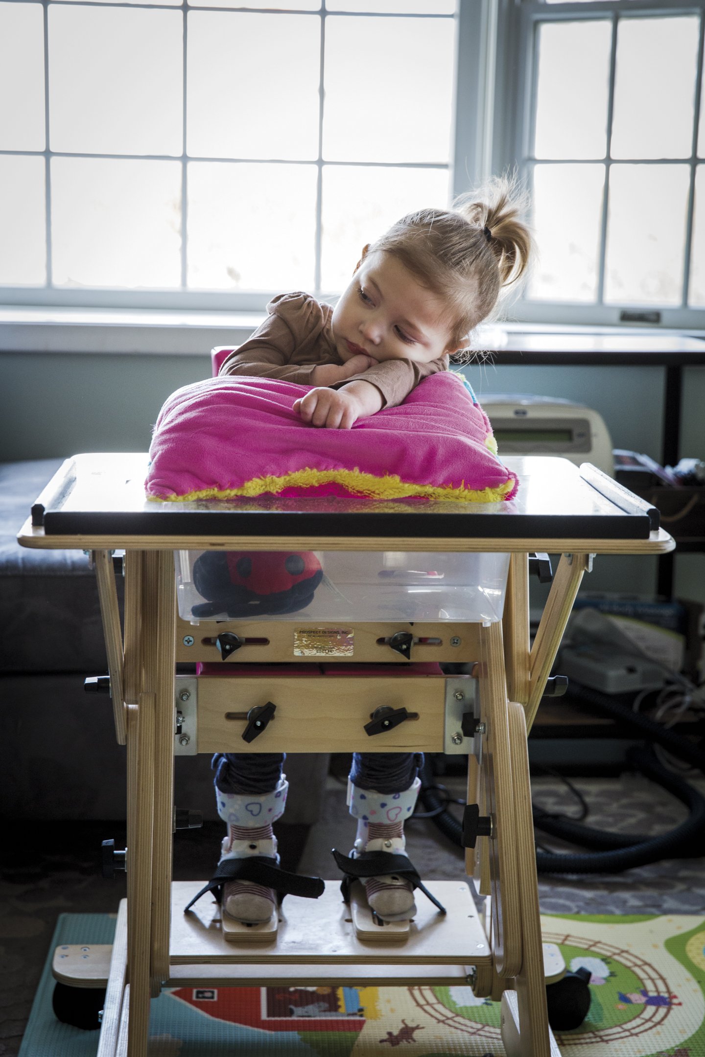 A young girl with blonde hair in a ponytail resting her chin on a pink pillow on a standing desk, with her feet in foot braces on a standing frame, inside a room with large window panes.