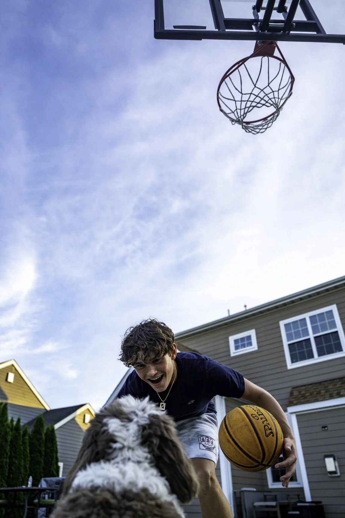 A young man playing basketball outside, holding an orange basketball, and smiling while interacting with a fluffy dog in a backyard with houses and a blue sky in the background.