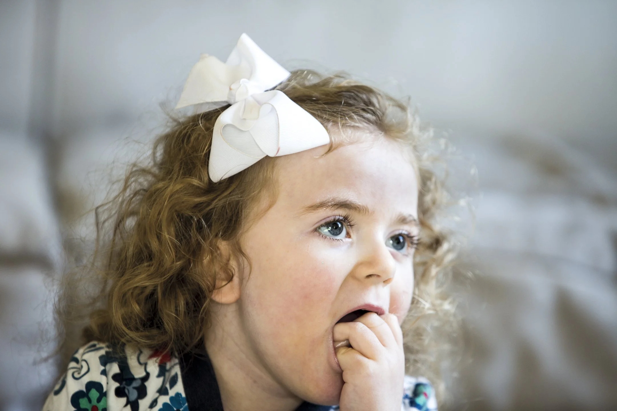 A young girl with curly hair and a large white bow on her head, sitting indoors with her hand near her mouth, appearing surprised or worried.
