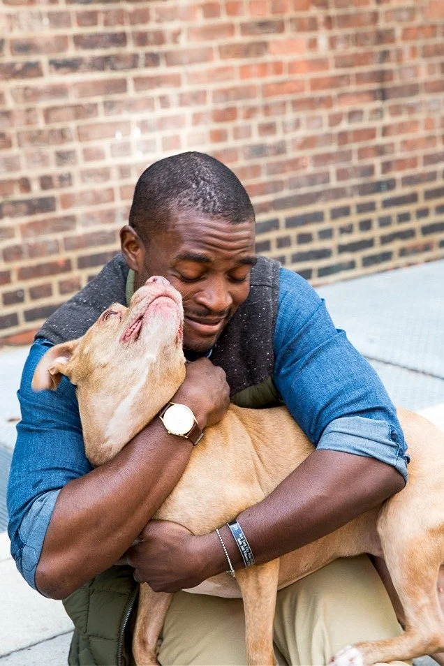A man hugging a yellow dog with his eyes closed, showing affection, against a brick wall background.