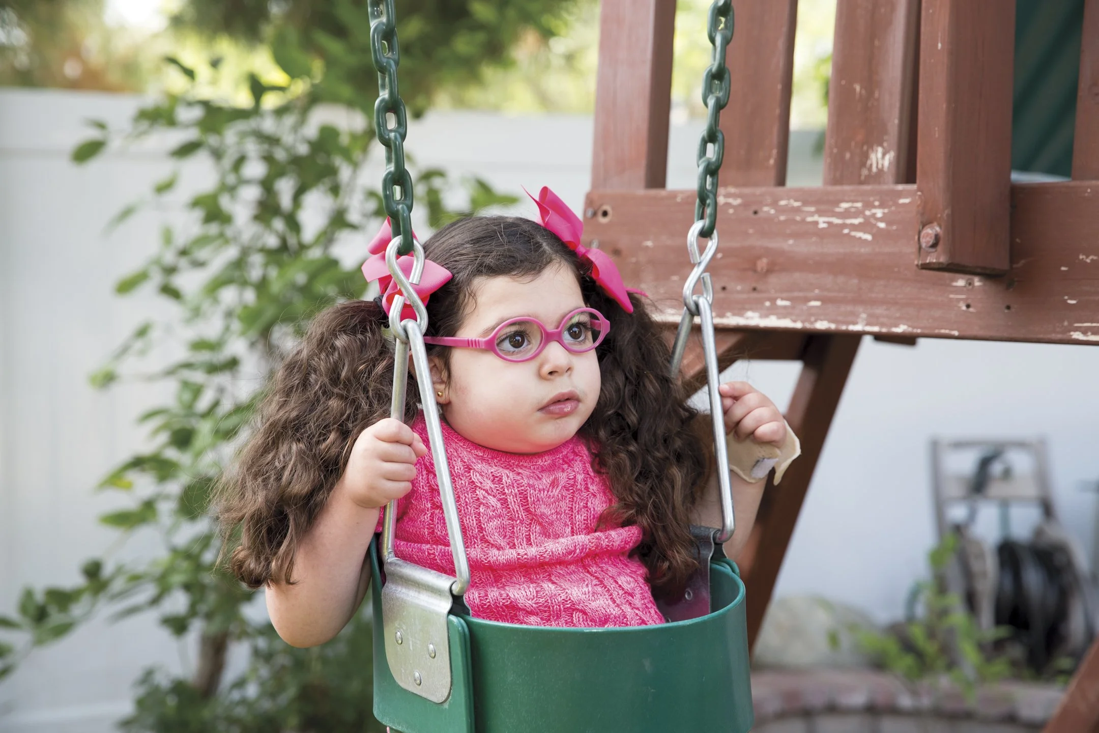 A young girl with curly hair in pigtails, wearing pink glasses and a pink shirt, sitting in a green baby swing on a wooden outdoor play structure.