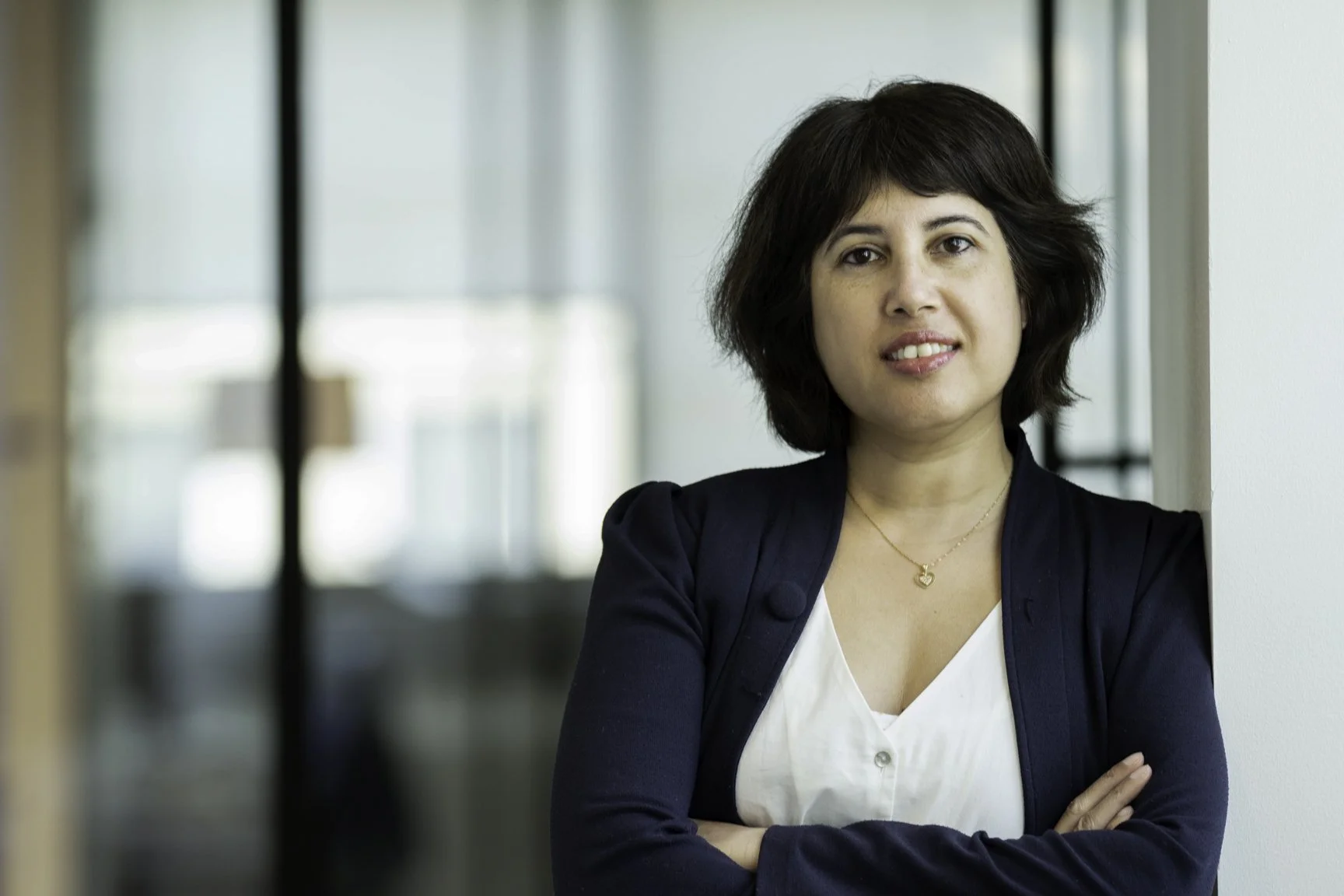 A woman with short dark hair and a white top, wearing a navy blazer and a gold necklace, standing with her arms crossed next to a white wall in an office setting.