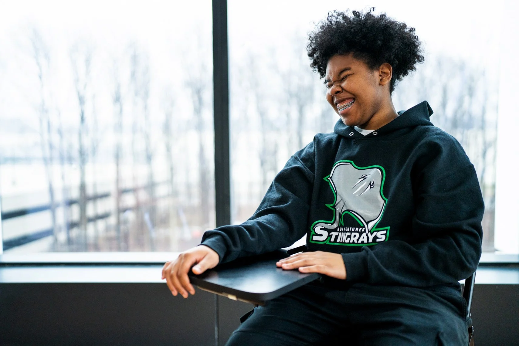 Young woman laughing while sitting near a window, wearing a black hoodie with a sports team logo and holding a clipboard.