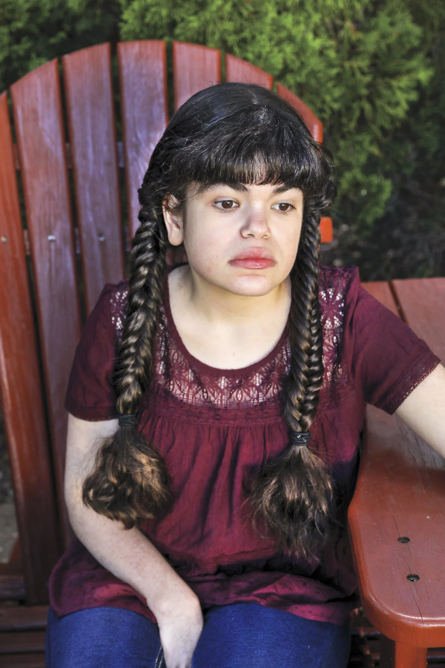 A young girl with dark braided hair wearing a maroon top, sitting on a wooden outdoor bench with greenery in the background.