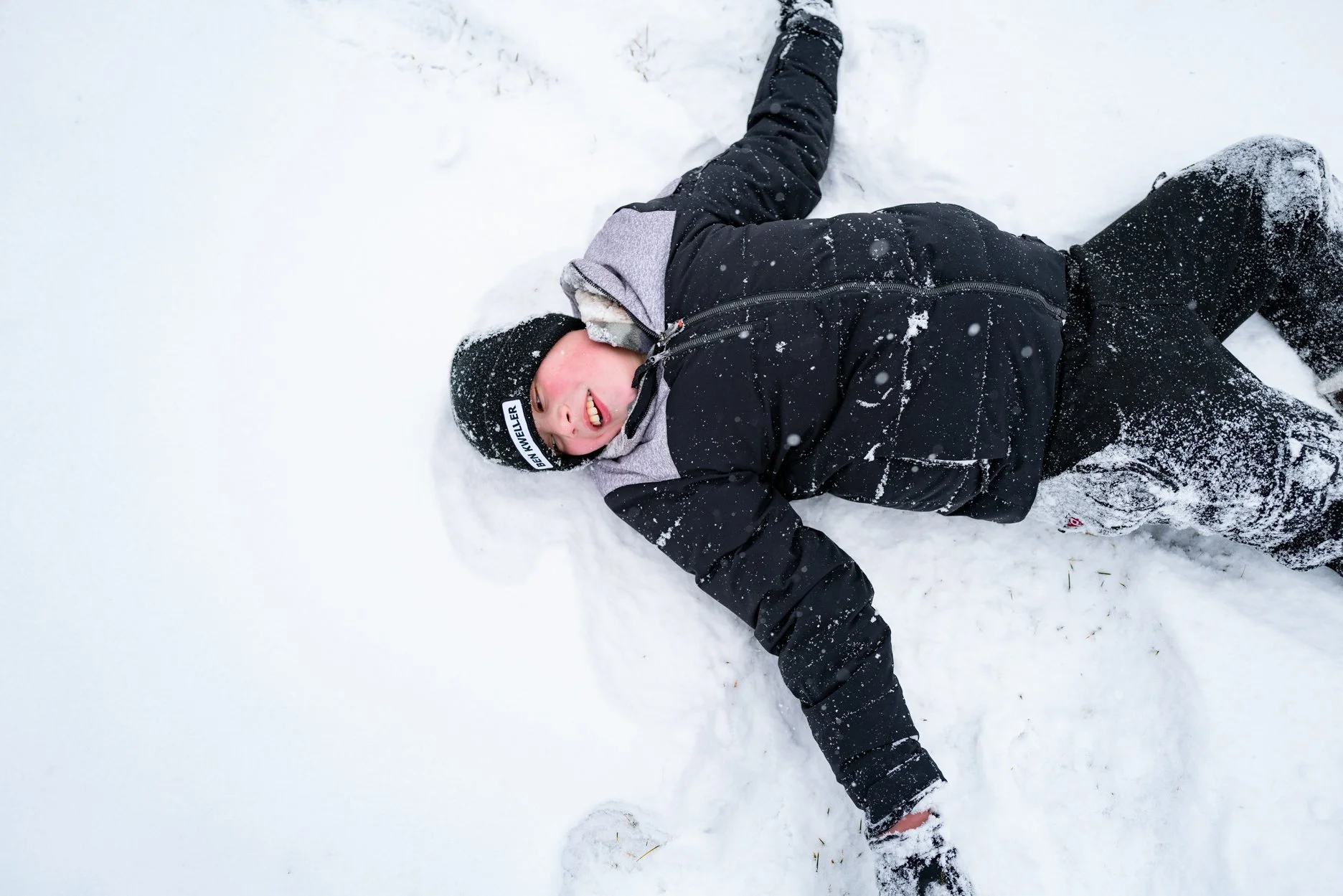 Child lying on snow, smiling, wearing black and gray winter jacket, black pants, and black beanie, making a snow angel.