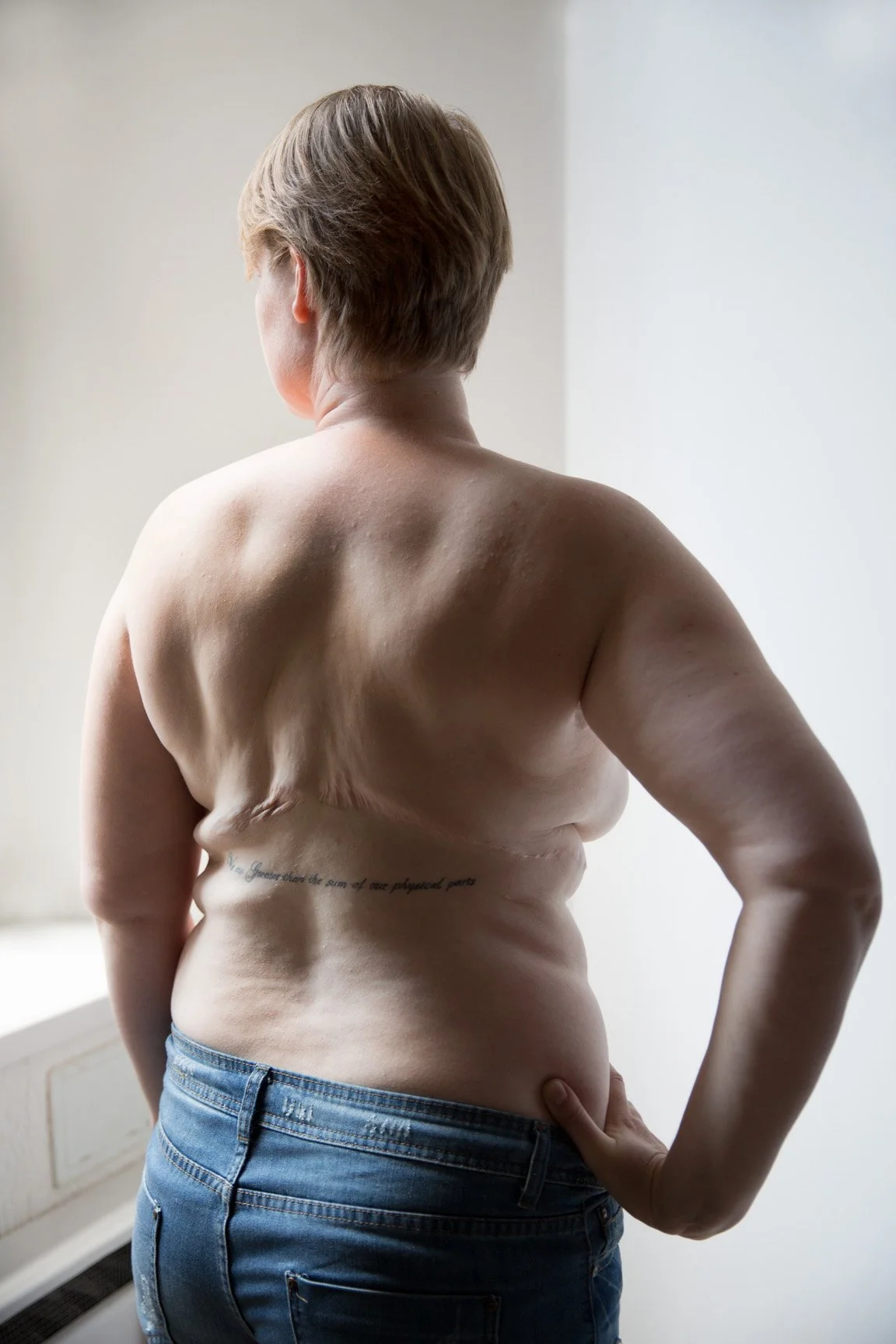 Back view of a topless person with short hair, standing near a window with a tattoo on their side.