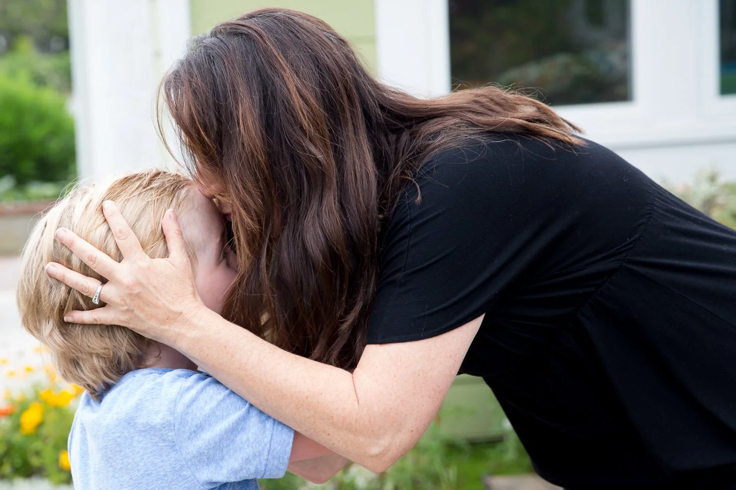 A woman with long dark hair gently holds a young boy with blonde hair, leaning in for a kiss outside near a house with a flower garden.