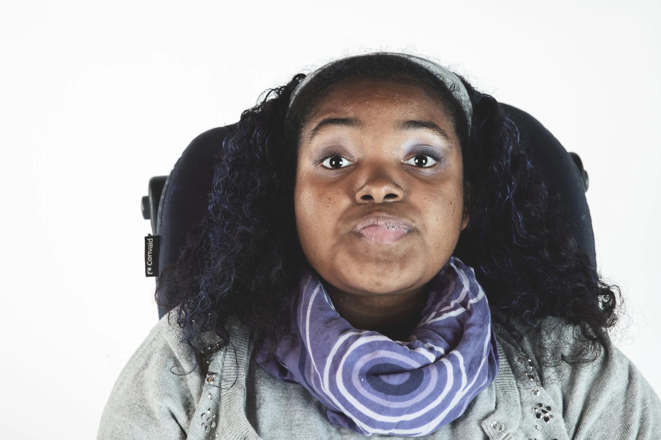 Close-up of a woman with dark skin and curly black hair, sitting in a wheelchair, wearing a gray jacket and a purple striped scarf, against a plain white background.