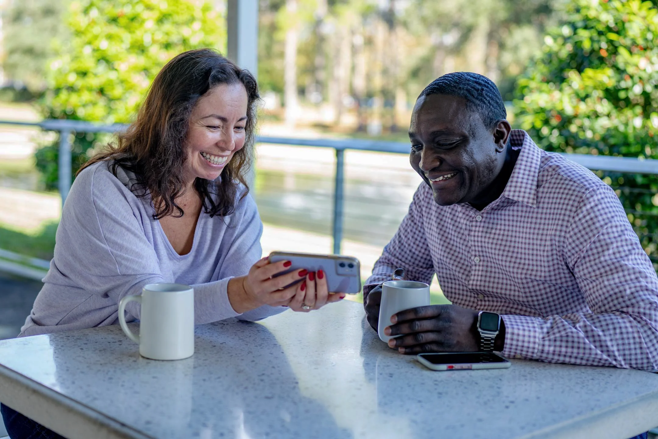 A woman and a man sitting at an outdoor table, smiling while looking at a smartphone.