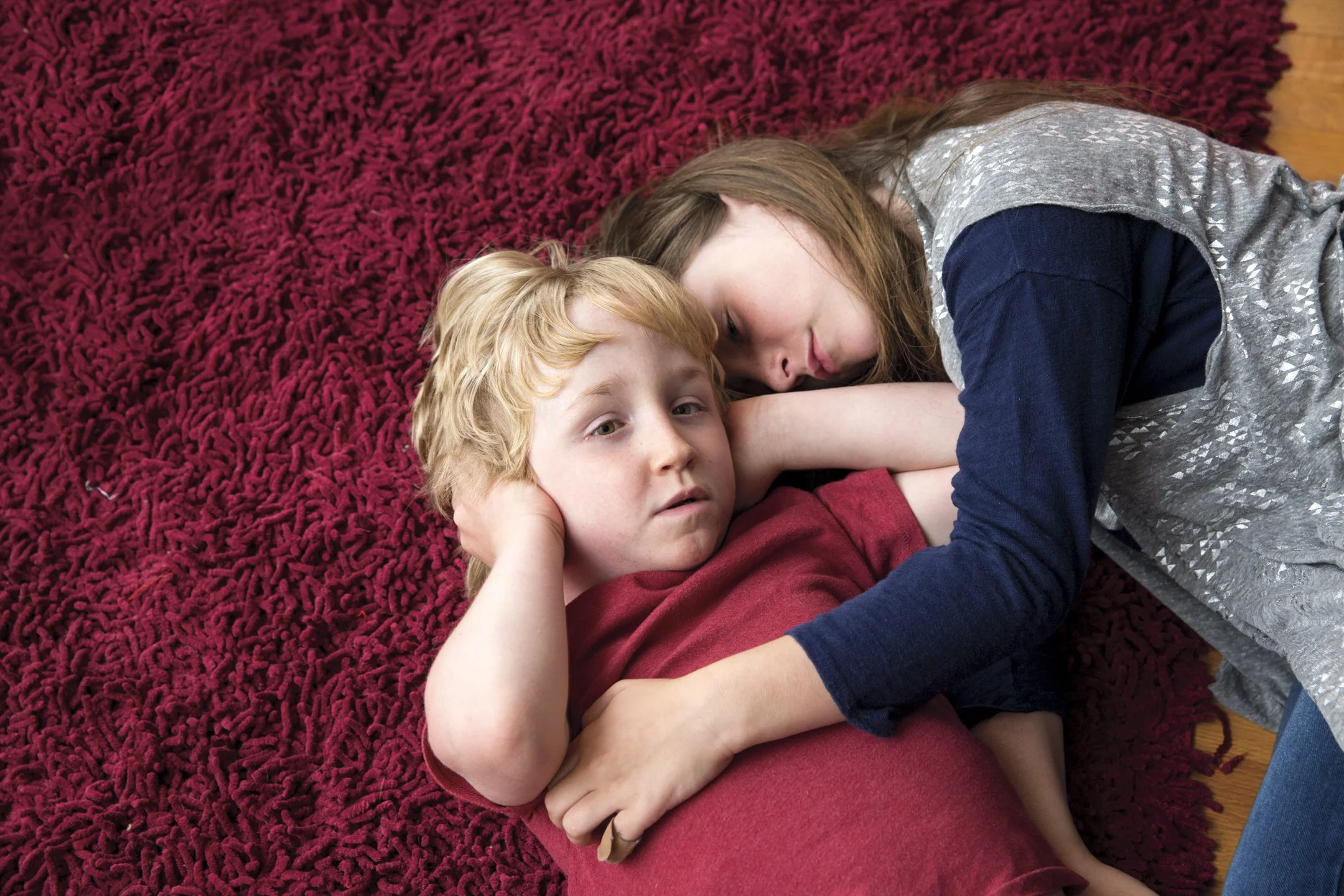 A young girl cuddling with a boy while lying on a red shag rug.