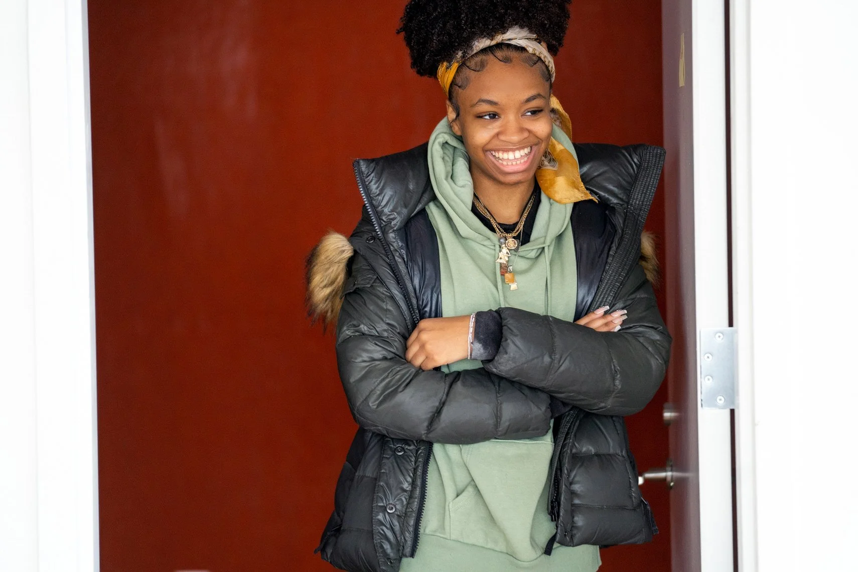 Young woman with dark curly hair in an afro, wearing a yellow headscarf, a black puffer jacket with fur trim, and a green hoodie, standing at a doorway with her arms crossed, smiling.