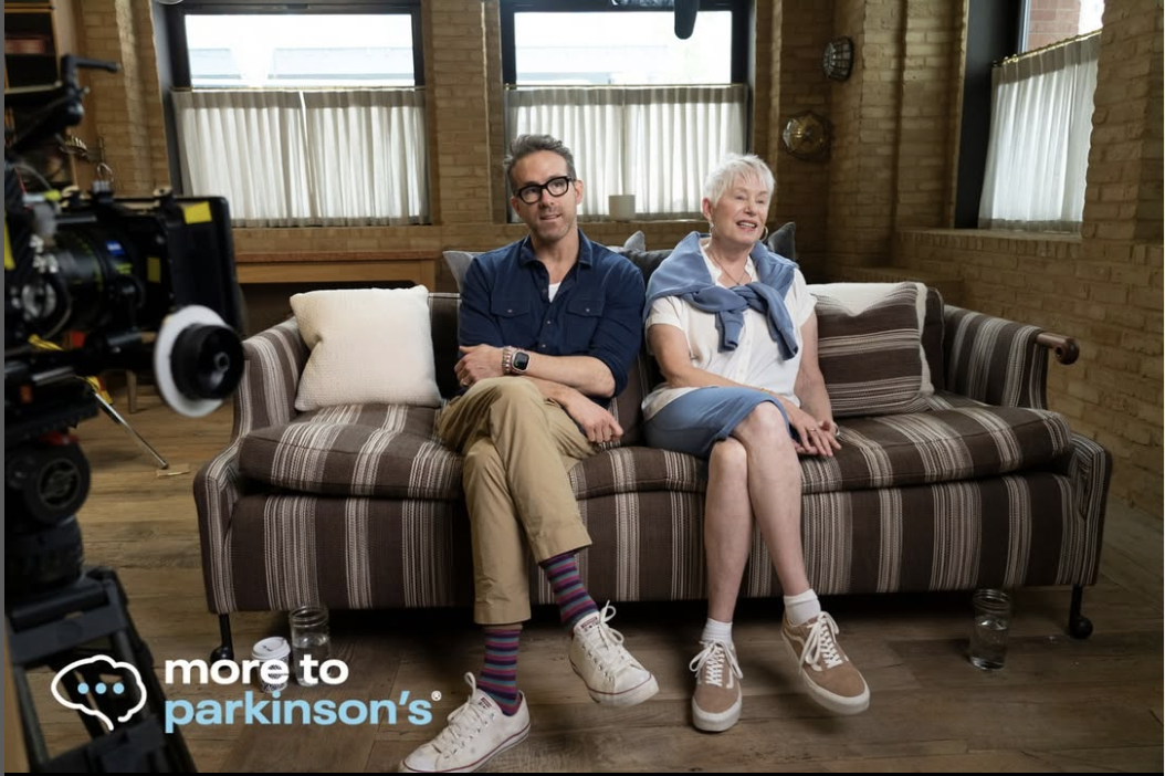 A young man and an older woman sitting on a striped sofa in a brightly lit room with brick walls and large windows, during a video shoot for More to Parkinson's.