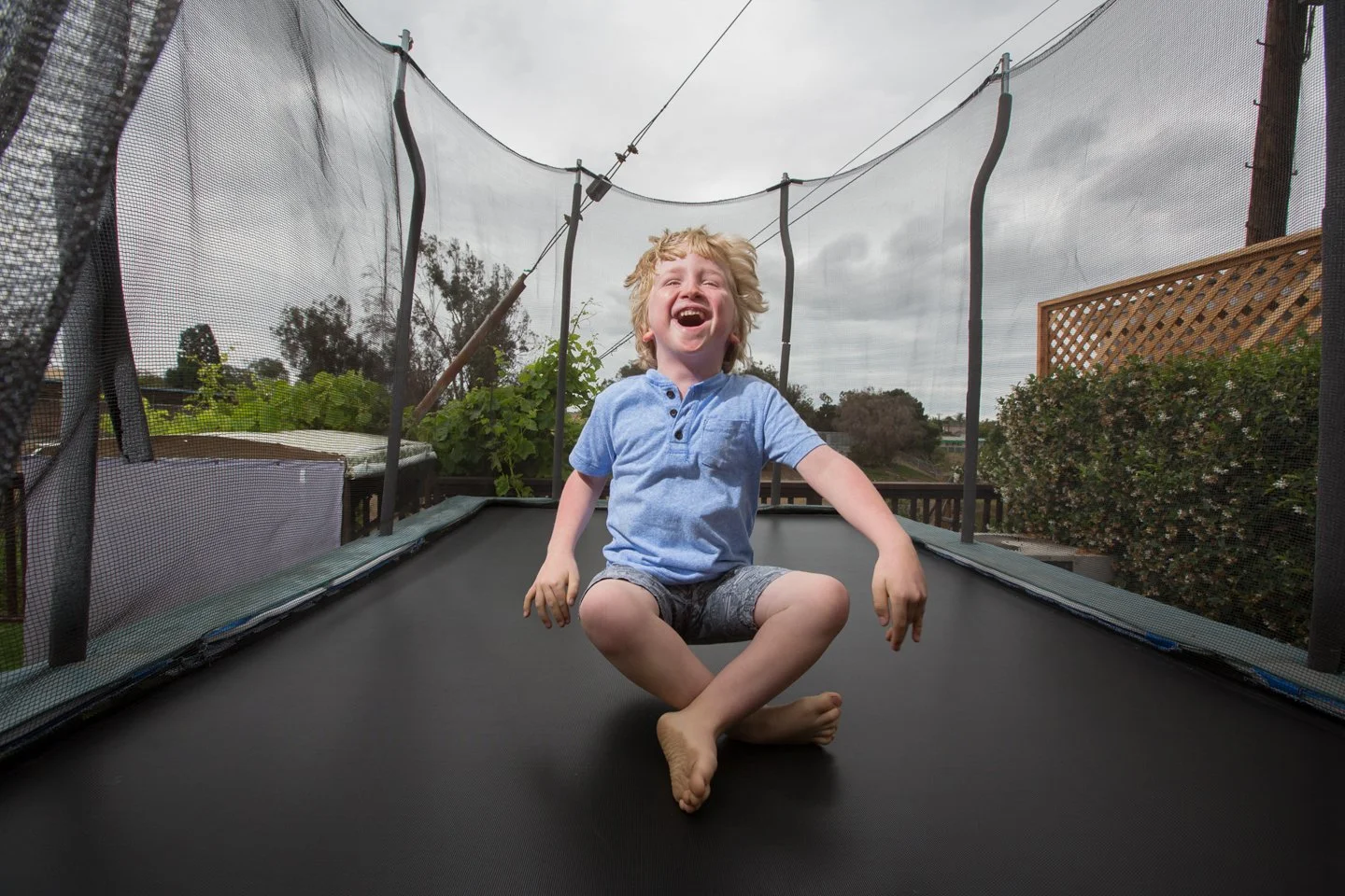 A young boy with blond hair wearing a blue shirt and shorts happily laughing while kneeling on a trampoline outdoors, with a cloudy sky and trees in the background.