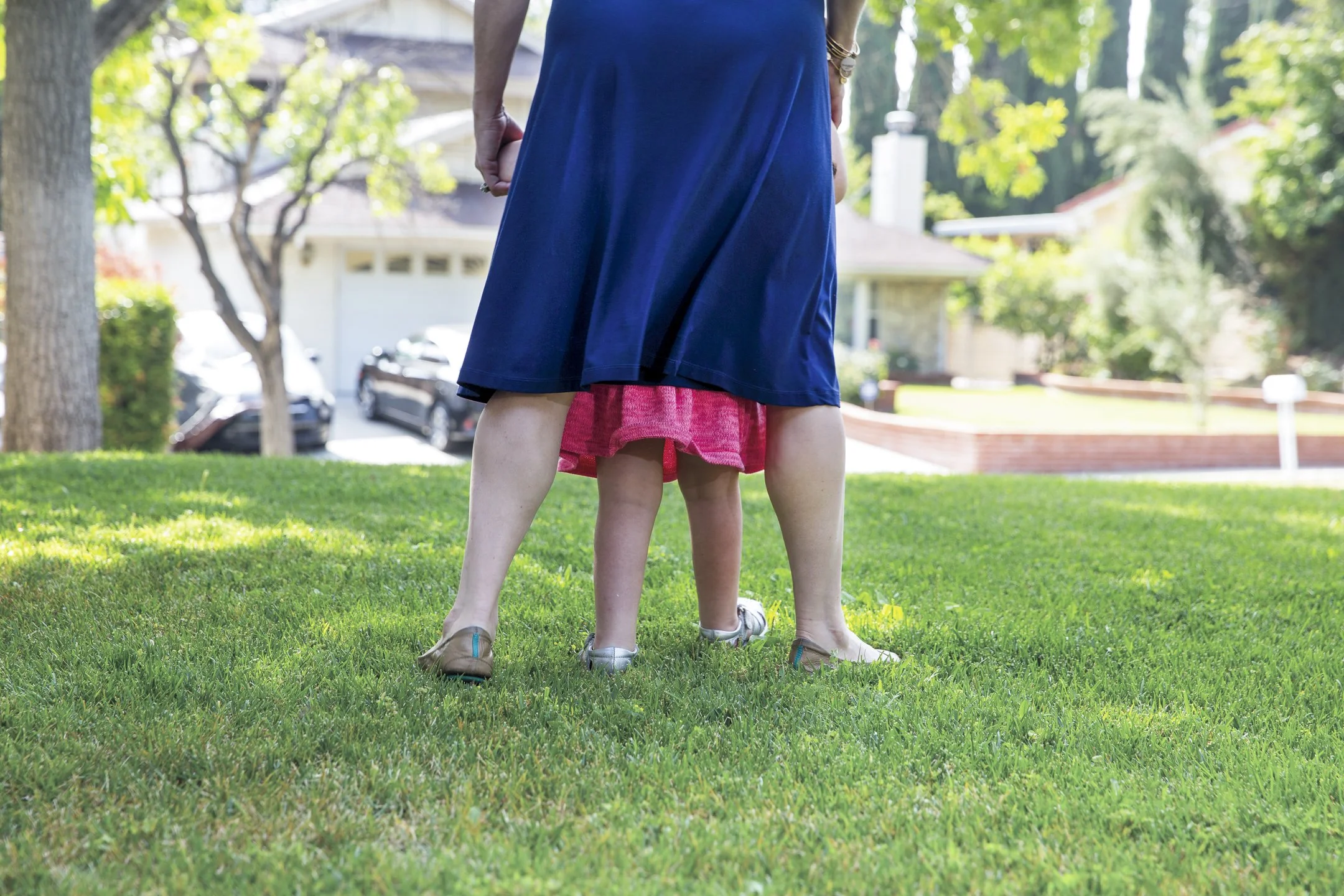 Adult and child standing on a grassy lawn, with the child's head partially obscured by the adult's dress, in a suburban neighborhood.