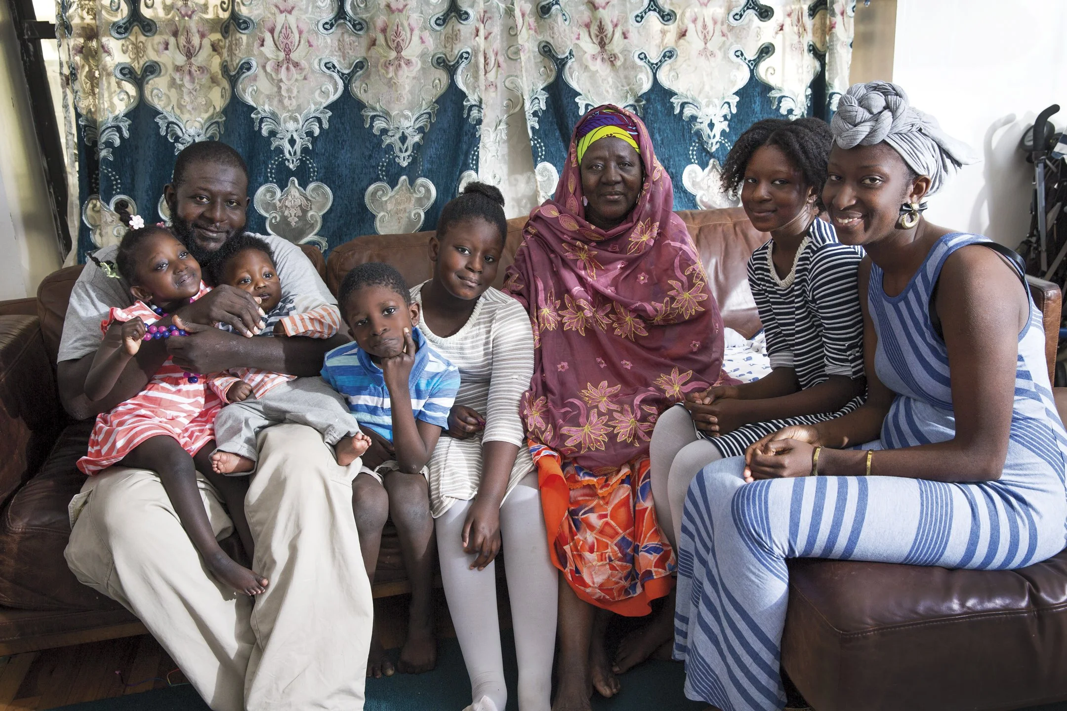 Family portrait of eight people sitting on a brown leather sofa indoors, with a blue and white patterned curtain in the background. The group includes children, a man, a woman, and an older woman, all smiling and dressed casually.
