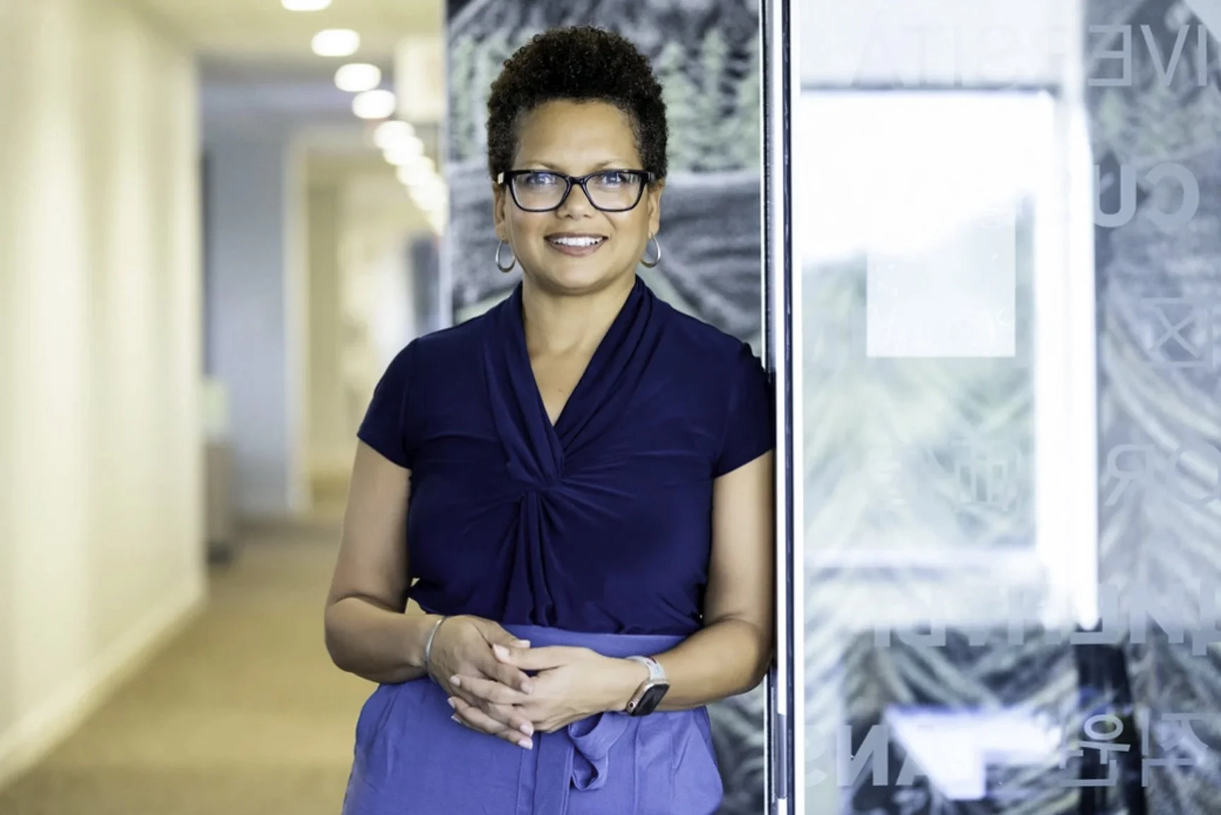 A middle-aged woman with short curly hair, glasses, and hoop earrings, wearing a navy blue top and a blue skirt, standing in a modern indoor space near an elevator with her hands clasped.