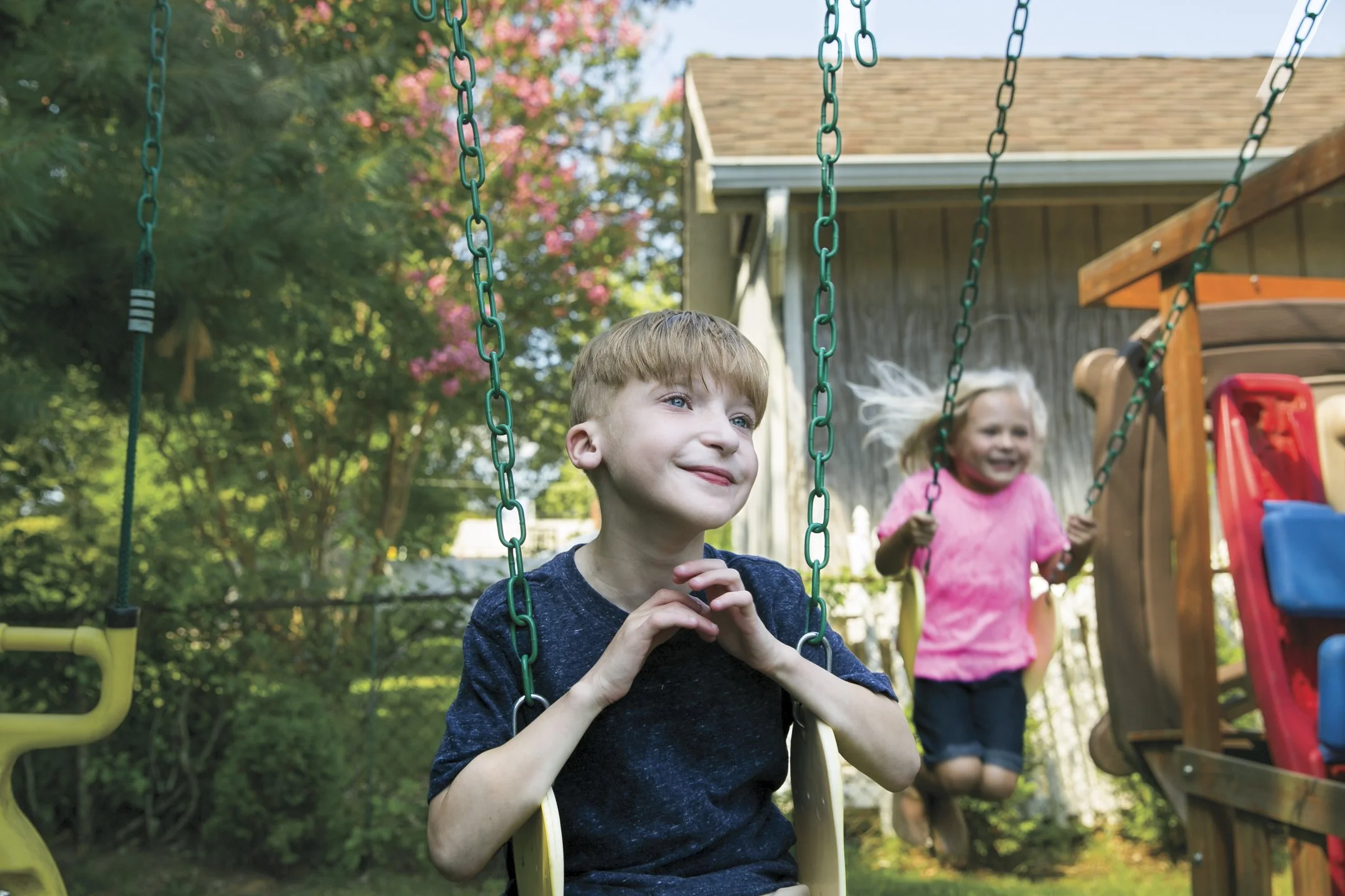 Two children, a boy and a girl, playing on swings in a backyard with trees and a shed.