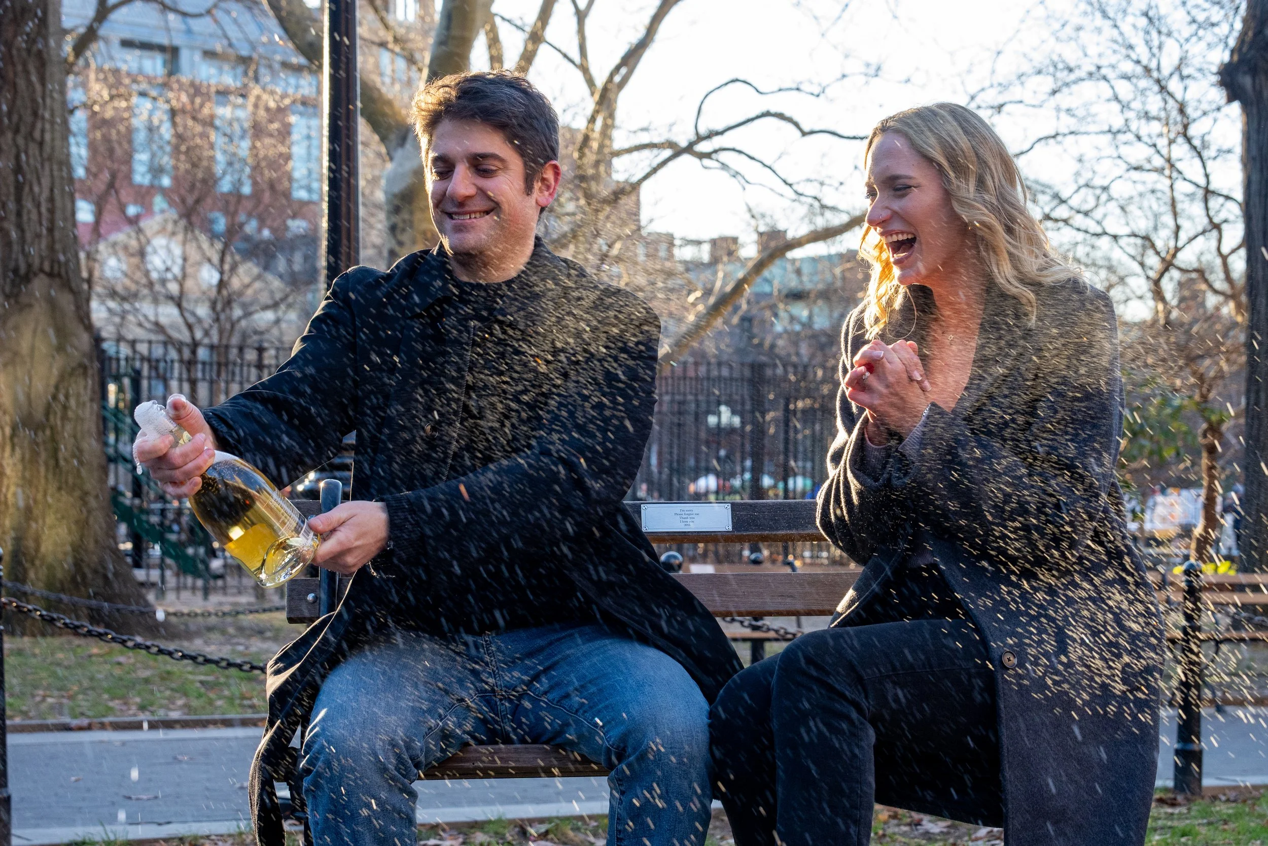A man and woman sitting on a park bench celebrating, with the man opening a bottle of champagne and champagne spray in the air, both smiling and happy.