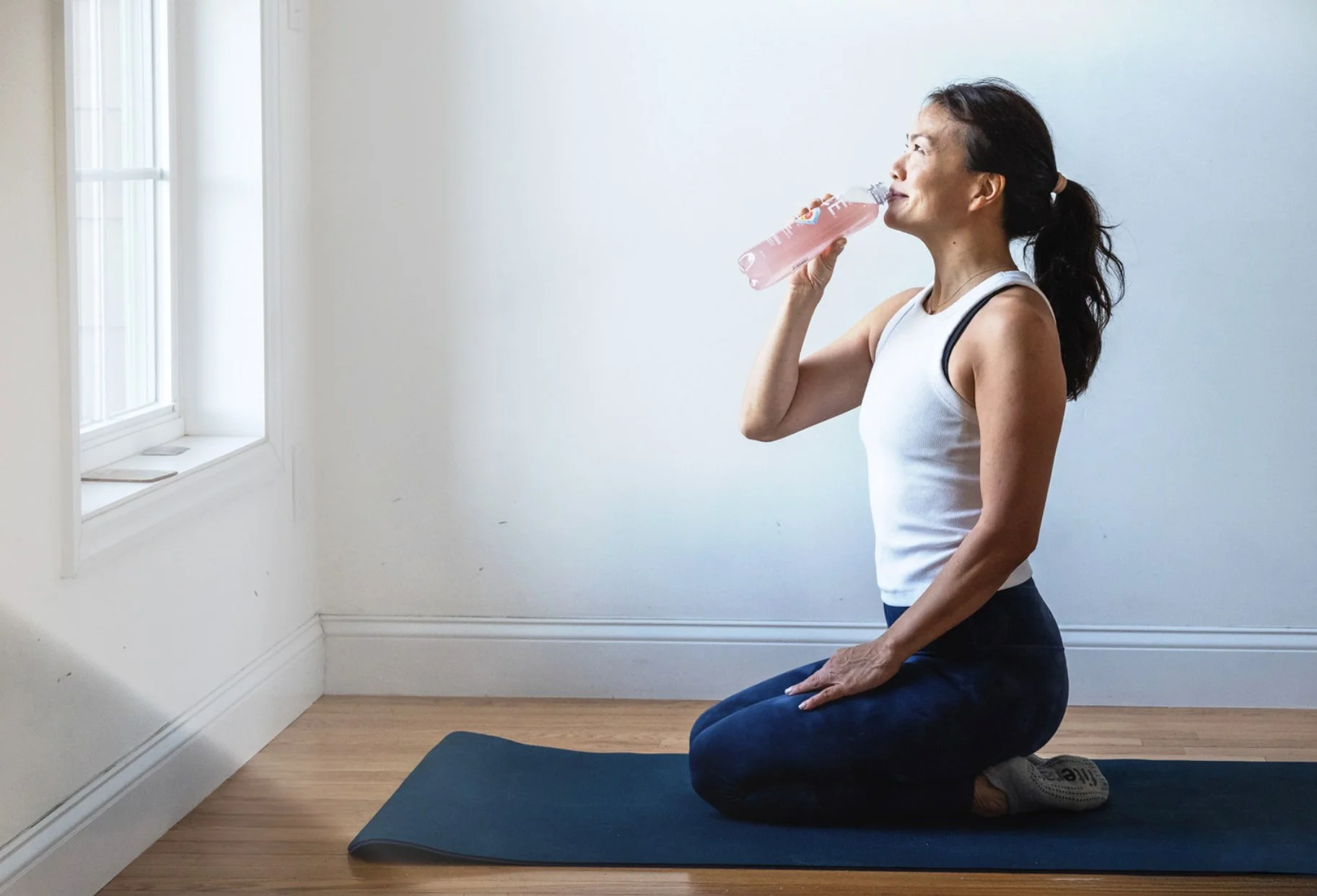 A woman kneeling on a yoga mat near a window, drinking water from a pink water bottle.
