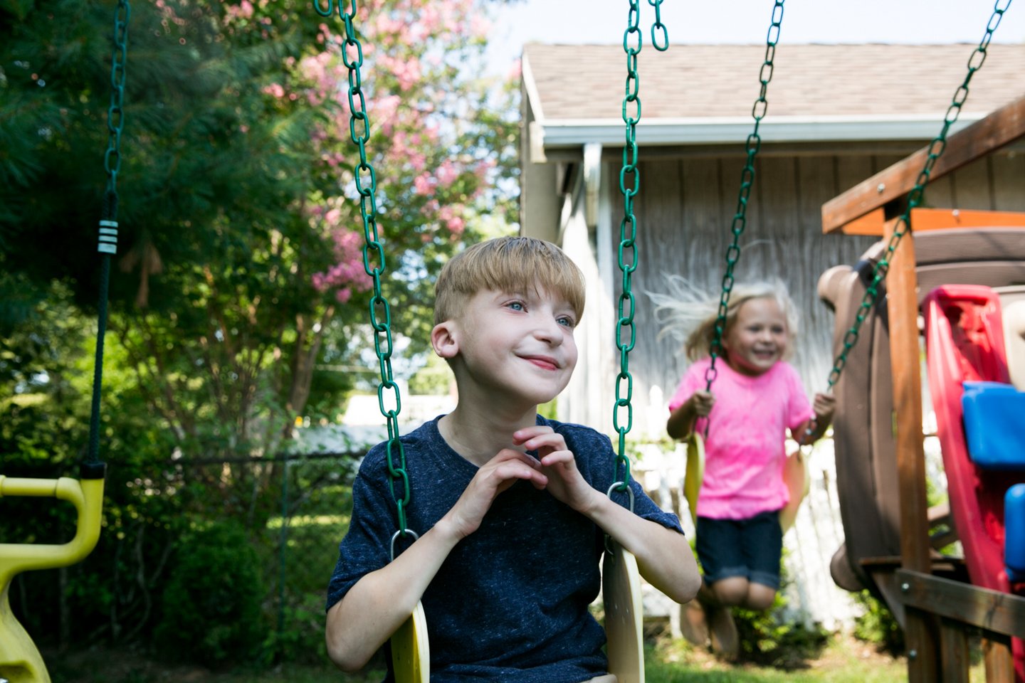 A young boy with blonde hair and blue eyes on a swing in a backyard, smiling and looking content. A young girl with blonde hair in pigtails wearing a pink shirt and dark shorts is in the background also on a swing, smiling and having fun.