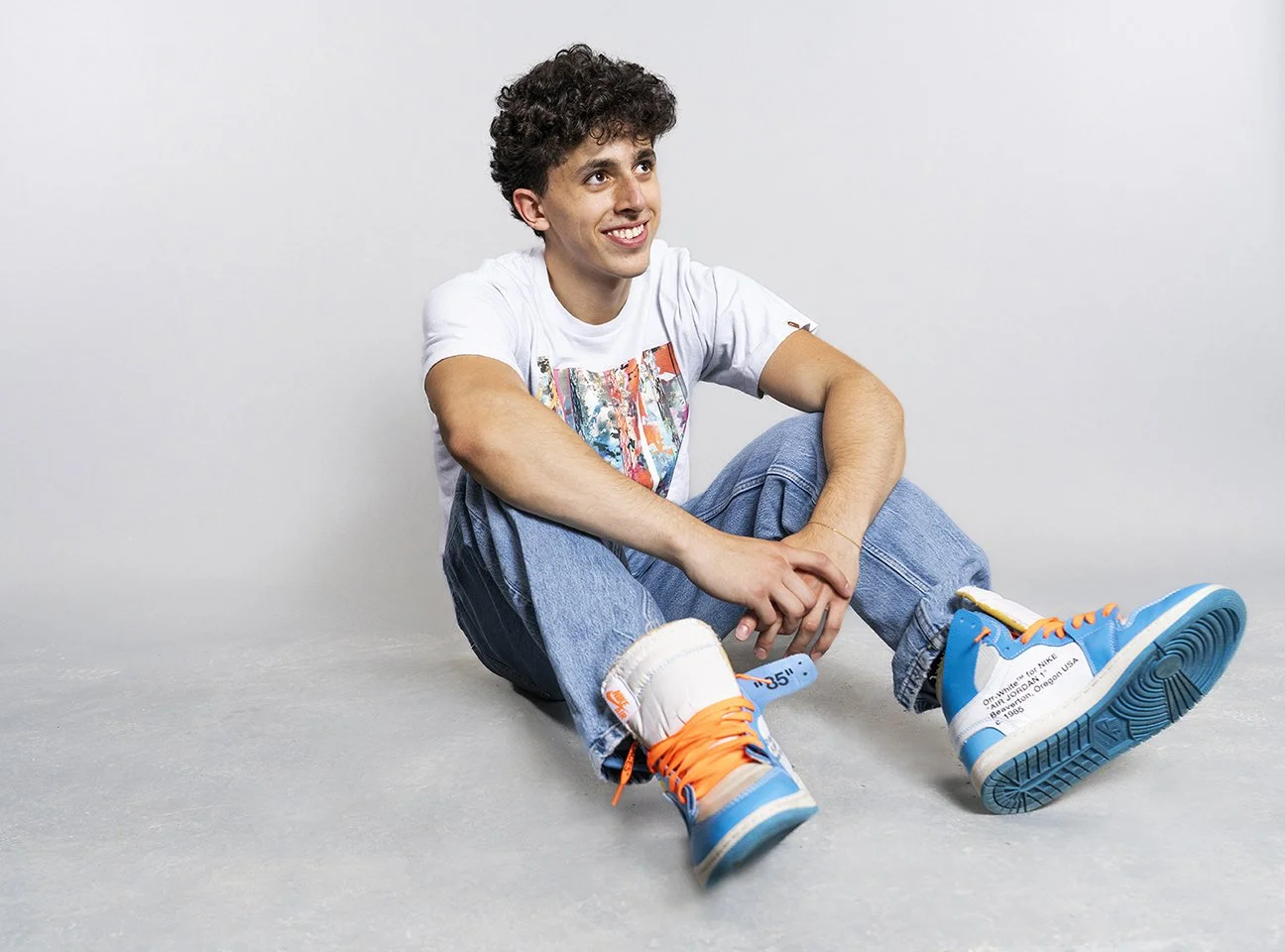 A young man with curly brown hair sitting on the floor, smiling, wearing a white t-shirt with colorful print, blue jeans, and bright blue and white sneakers with orange laces.