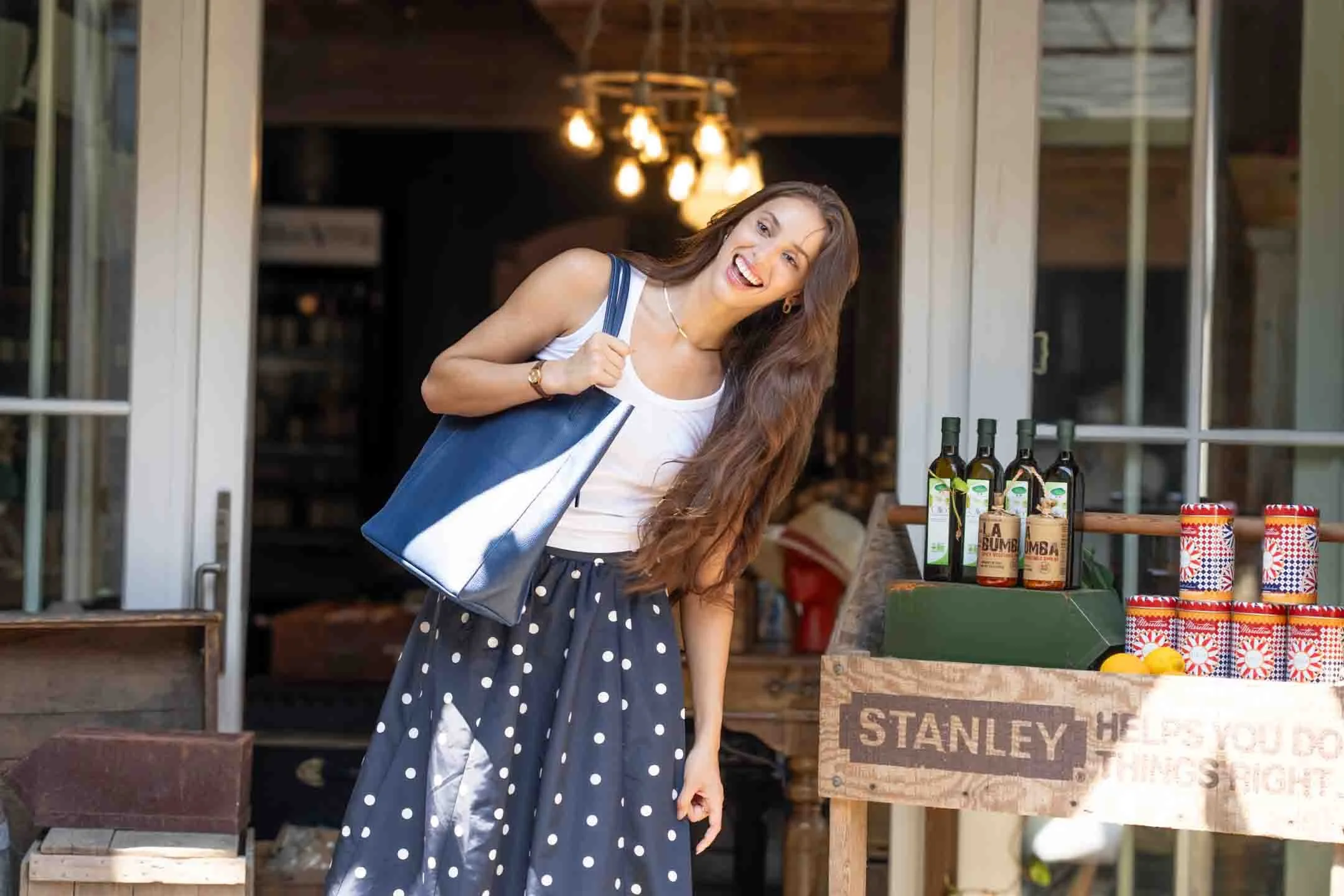A woman smiling and carrying a blue shopping bag, standing outside a store selling bottles and cans of food or drink.