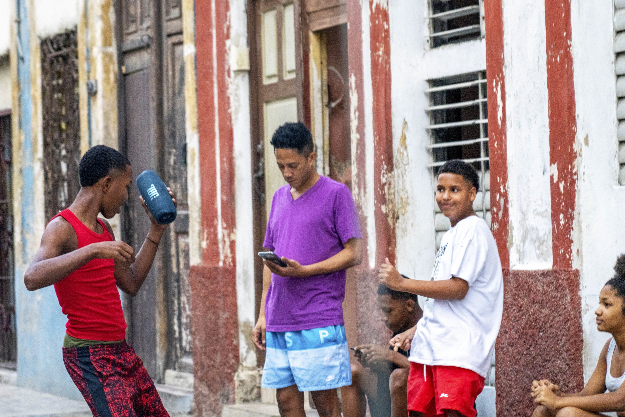 A group of young boys and a woman standing outside a colorful, weathered building, with some boys using mobile phones and one boy dancing.