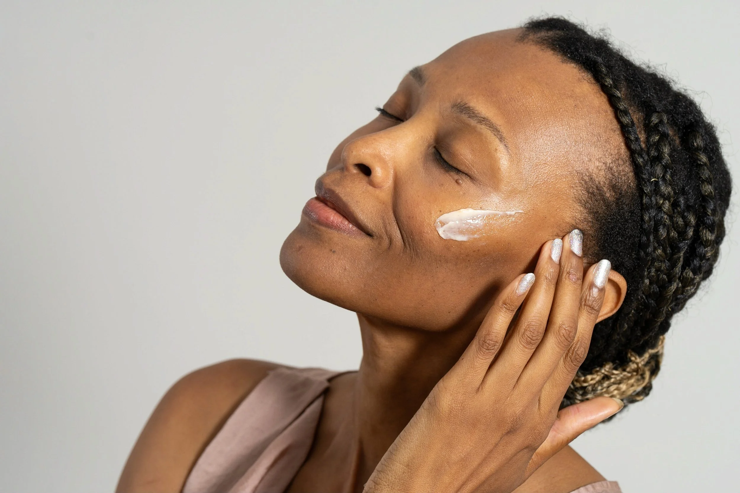 A woman applies skin moisturizer to her face with her eyes closed.