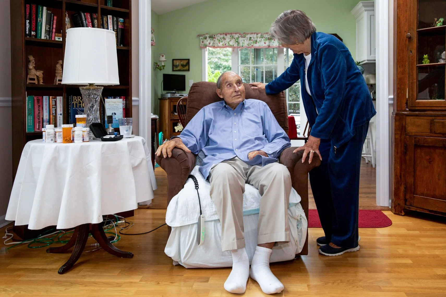 An elderly man with white socks is sitting in a recliner while a woman in blue pajamas leans towards him and speaks. The room has a bookshelf, a side table with medications, and a window with floral curtains.