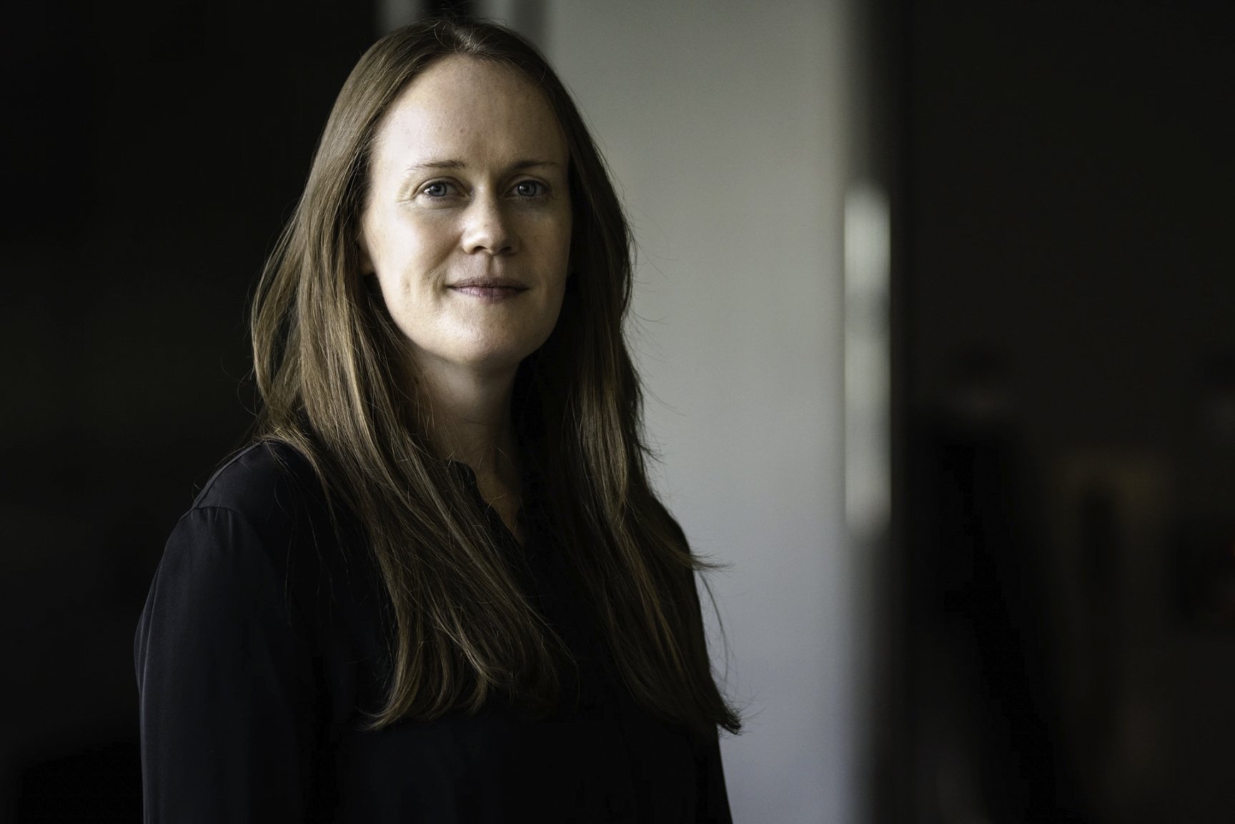 A woman with long brown hair and fair skin, wearing a black top, stands indoors against a blurred background with a light-colored wall on her right and a darker area on her left.