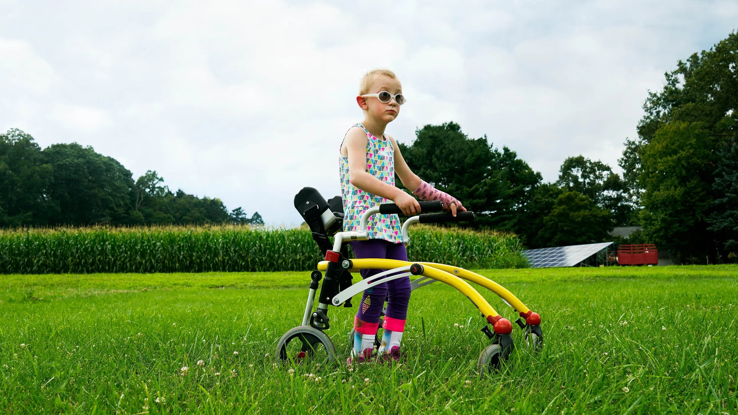 A young girl with sunglasses and colorful clothing standing on a walker outdoor on a grassy field with trees and a farm in the background.