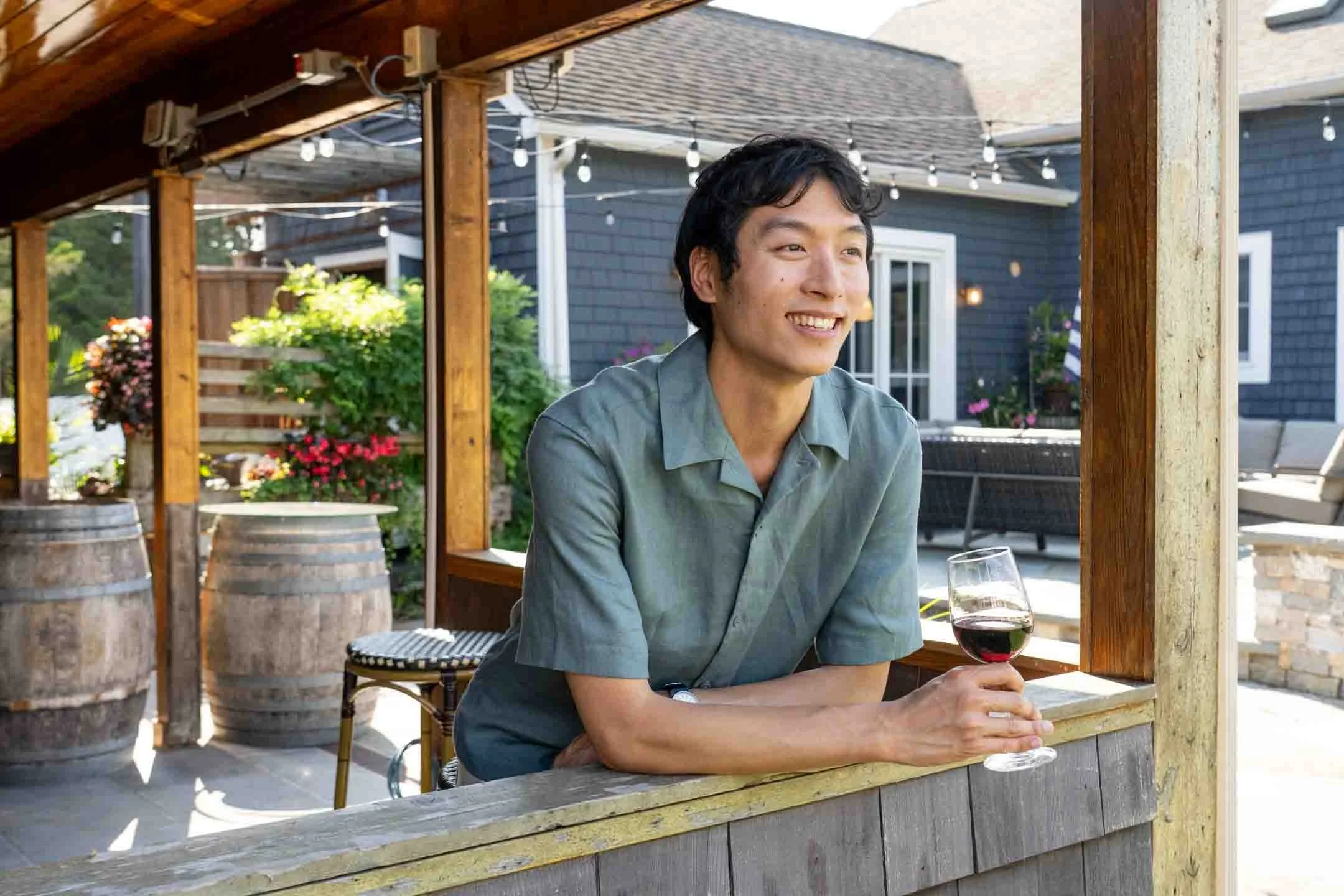 A smiling man with dark curly hair sitting outdoors at a bar, holding a glass of red wine. The setting appears to be a patio with wooden beams, barrel planters, and flowering plants, with a house in the background.