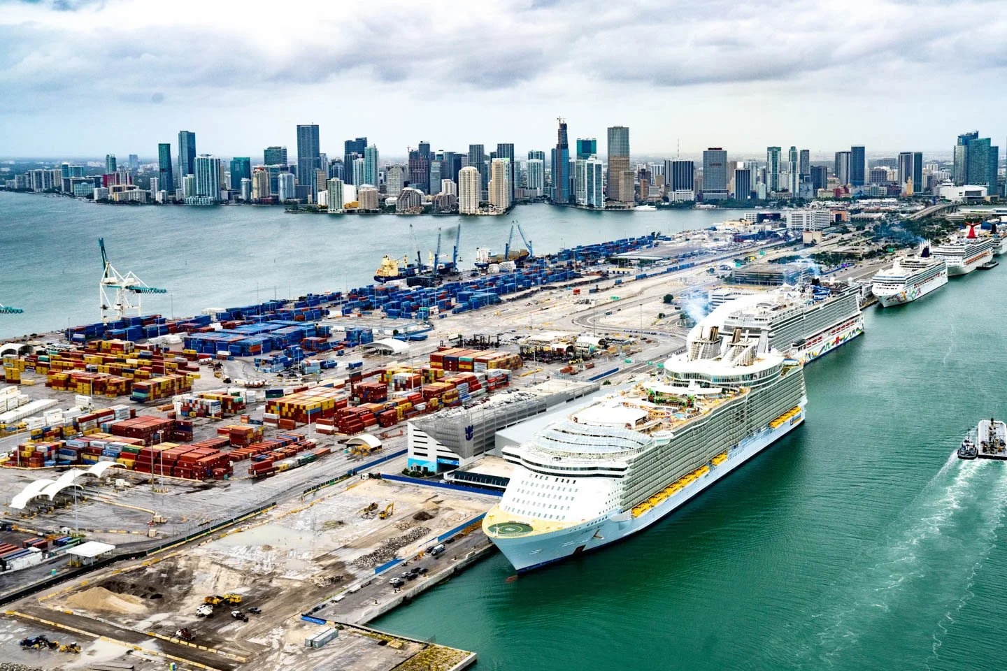 Aerial view of a busy port with cruise ships docked, colorful shipping containers, cranes, and a city skyline in the background.