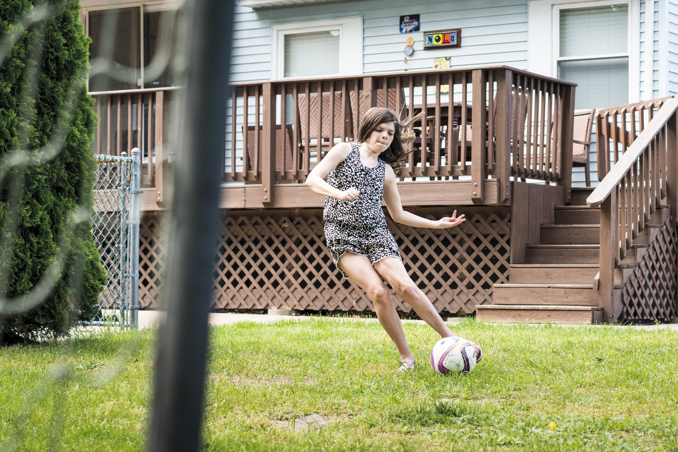 A young girl in a black and white patterned tank top and shorts playing with a soccer ball in a backyard with a wooden deck and stairs, partially obscured by a metal fence.
