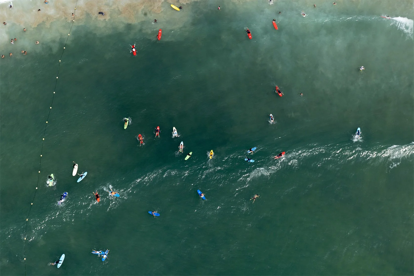 Aerial view of a crowded beach and ocean with people swimming and surfing, some near a yellow buoy line separating swimming area from surfing zone.