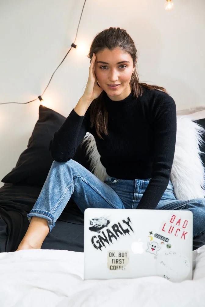 A young woman with long brown hair, wearing a black sweater and blue jeans, sitting on a bed with a laptop in front of her that has various stickers. She is resting her head on her hand and looking at the camera. The room has string lights on the wal