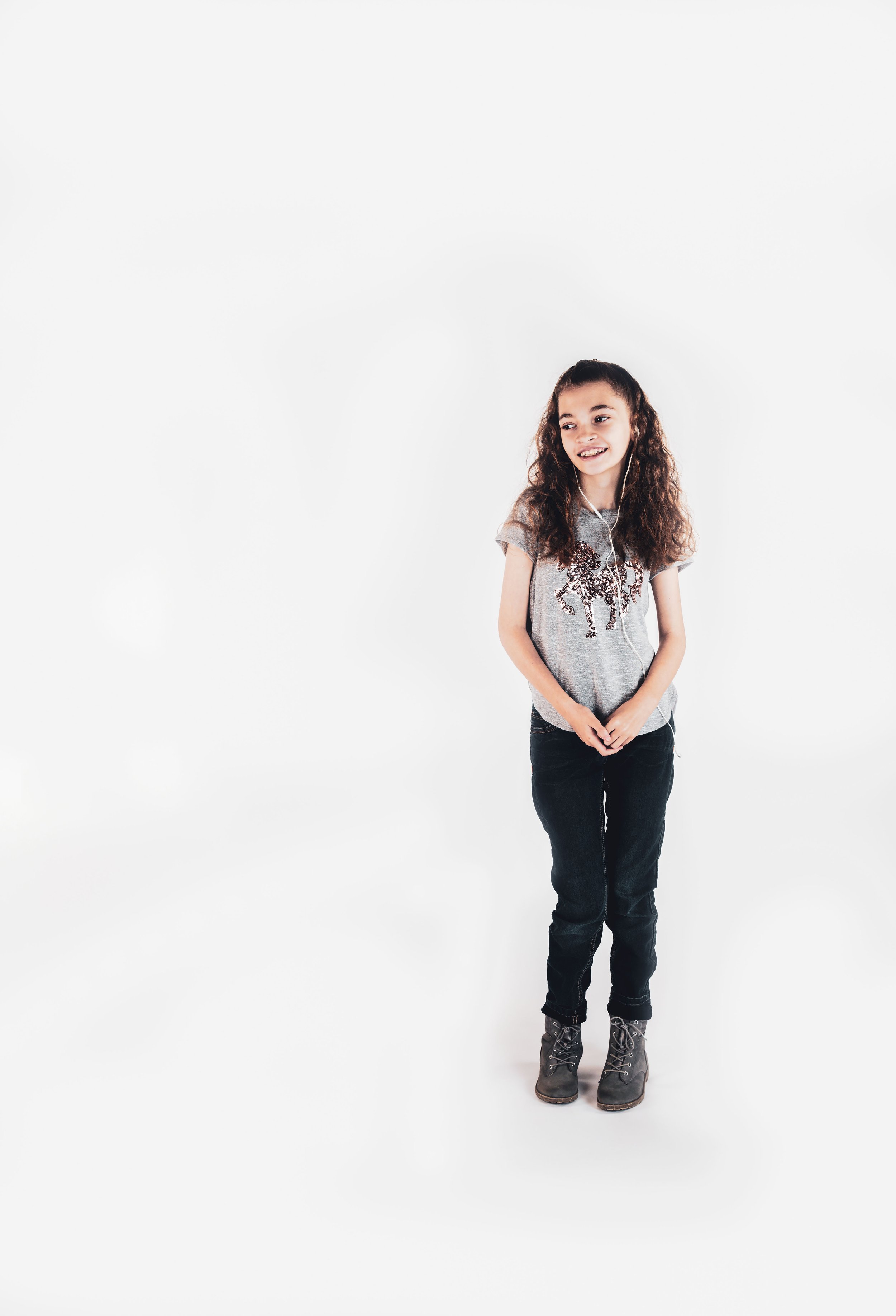 A young girl with curly brown hair wearing a grey t-shirt with a glittery animal design, black jeans, and grey boots, smiling and standing against a plain white background.