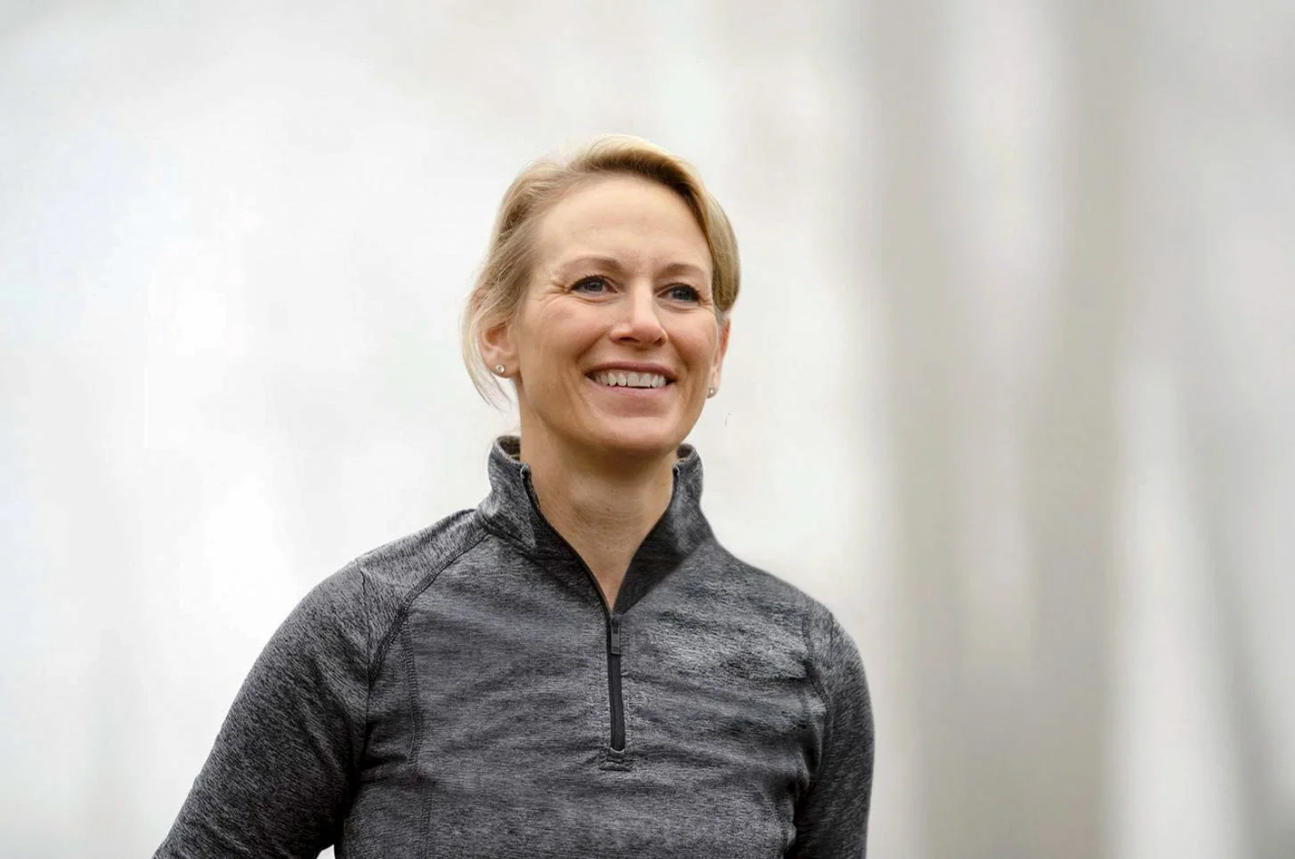 A smiling woman with short blonde hair wearing a gray quarter-zip athletic top, standing against a plain light-colored background.