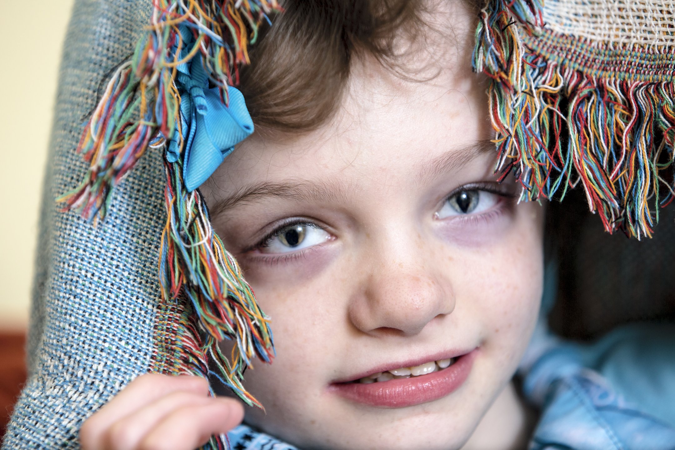 A young boy with blue eyes and freckles, wearing a colorful woven hat with fringe, holding it with one hand, looking into the camera with a slight smile.