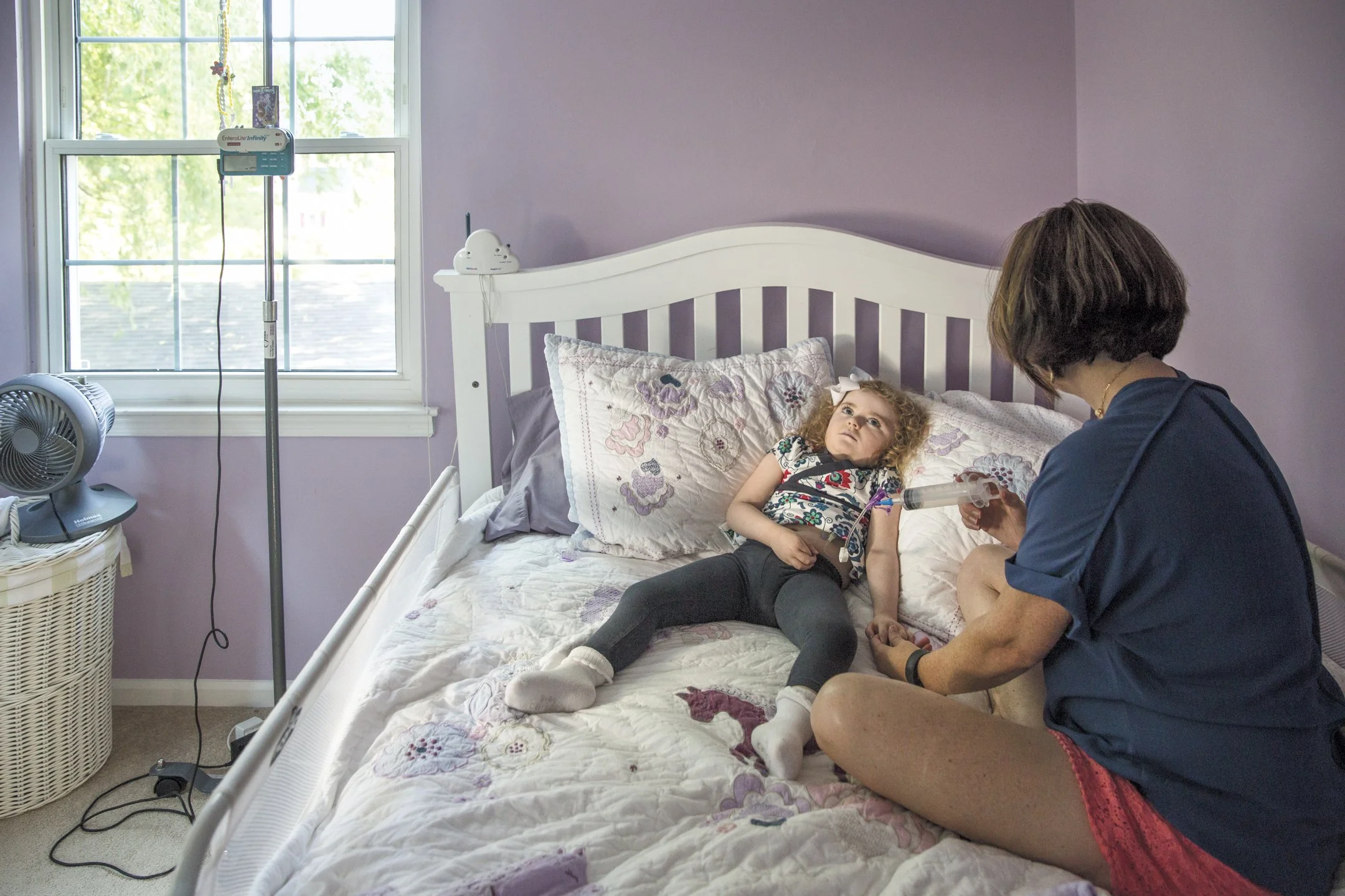 A young girl lying on a hospital bed connected to a nebulizer, with a woman sitting beside her administering treatment in a room with purple walls and a window.