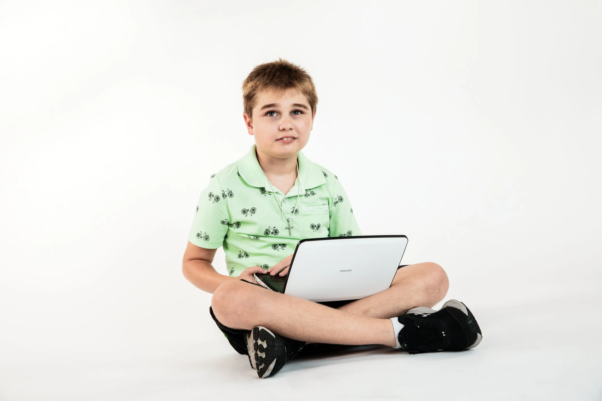 Young boy sitting cross-legged on the floor with a laptop, wearing a light green shirt with bicycle patterns, black shorts, and black sneakers, in front of a white background.