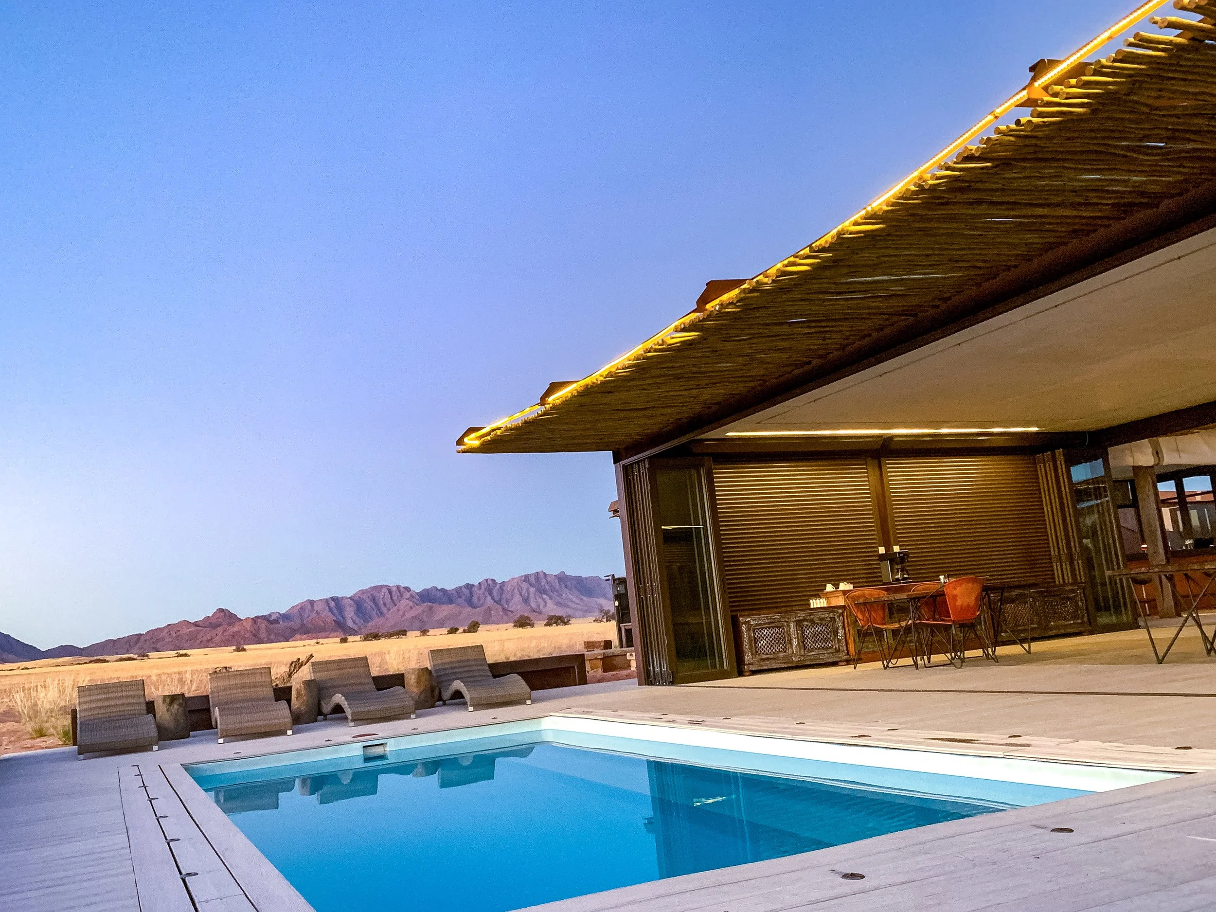 Luxury outdoor pool area at sunset with lounge chairs and a modern house with a deck featuring seating, overlooking desert mountains.