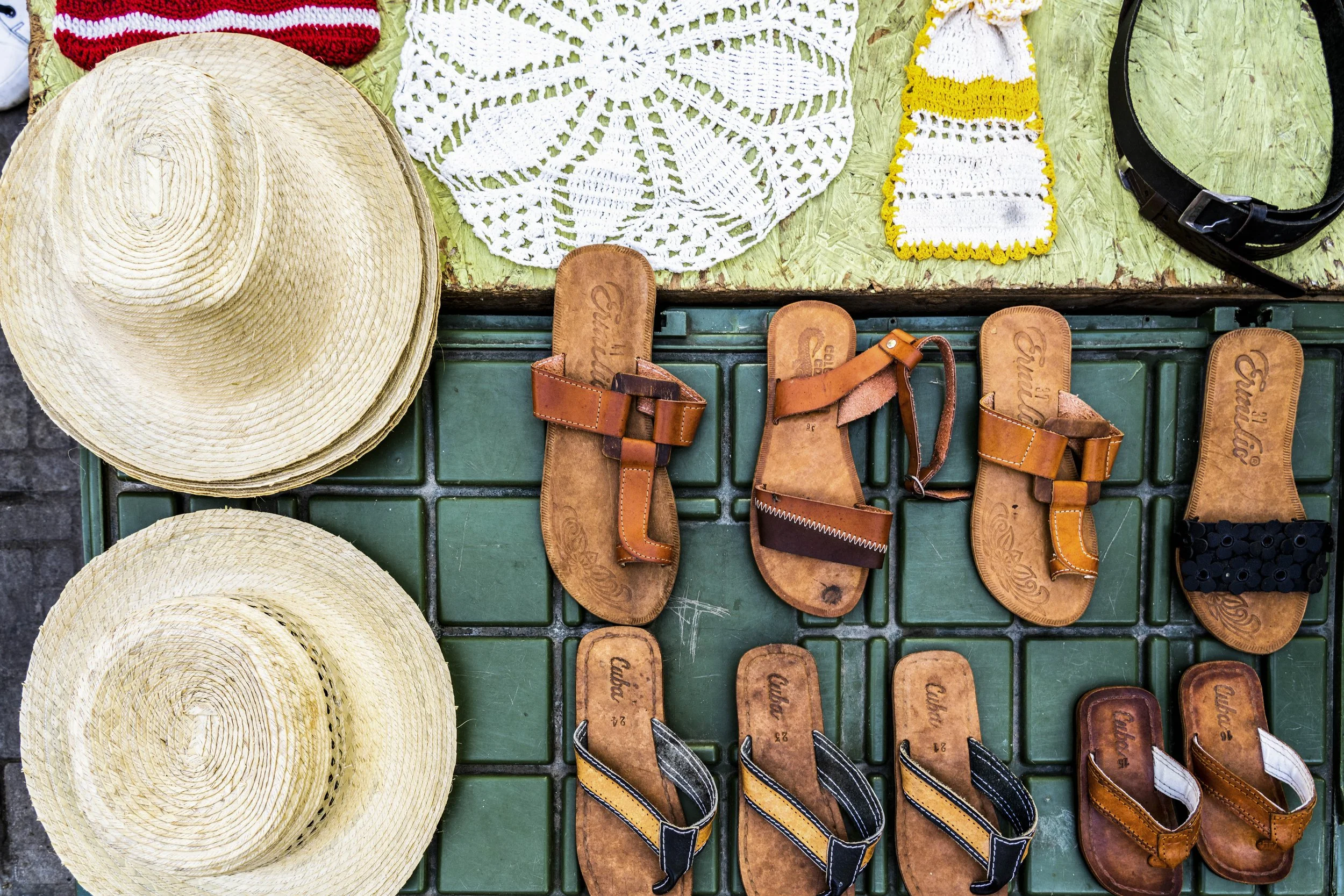 A display of tan sandals, straw hats, crocheted doilies, and a black belt on a table at a market