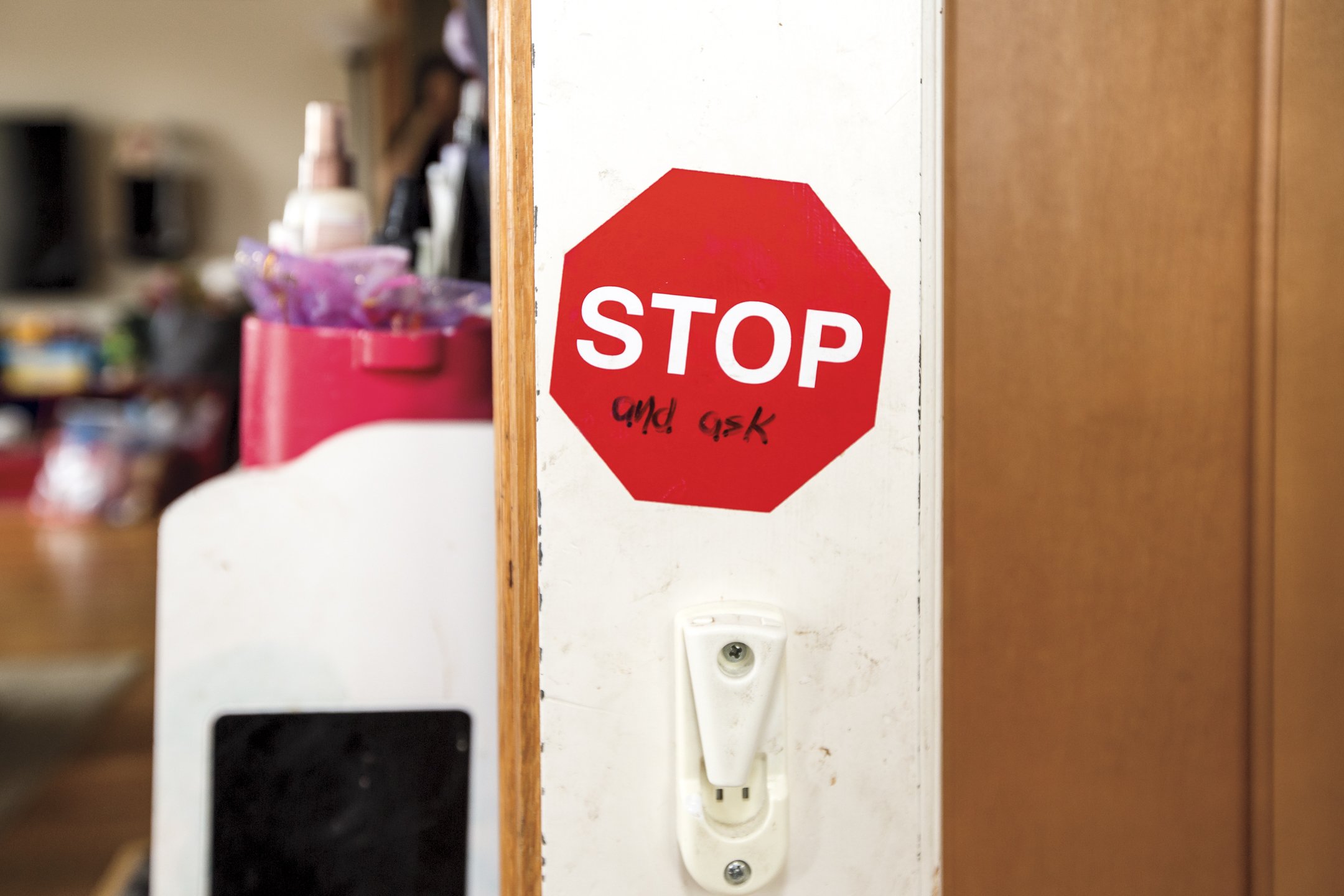 Red stop sign sticker with handwritten note underneath on a white wall, next to a power outlet, with a blurred background of a room with various objects.